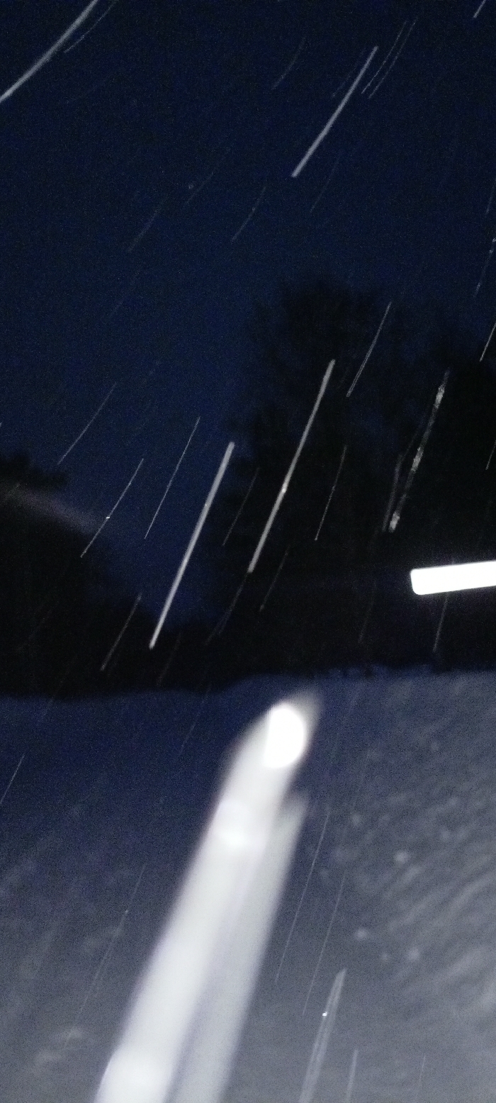 The image depicts a nighttime outdoor scene in Красный Маяк, Russia, characterized by active precipitation. The upper two-thirds of the frame show a dark, deep blue-black sky, with indistinct dark forms, possibly trees or distant structures, silhouetted against it. Numerous thin, bright white, diagonal streaks traverse the sky and mid-ground, indicating falling snow or rain illuminated by light sources and captured with motion blur. The bottom third of the image is a lighter, muted blue-grey surface, consistent with snow-covered ground or a wet road. A prominent, elongated bright white light source, extending from the bottom center and angling upwards to the left, dominates the foreground, showing significant motion blur and resembling a vehicle headlight or a strong reflection. A smaller, distinct white oval light is visible above and to the right of this primary light. Further to the right, near the center-right edge, a horizontal white rectangular light is present, suggesting a distant streetlamp or building light. No individuals or specific, clearly defined objects other than light sources and precipitation are discernible.