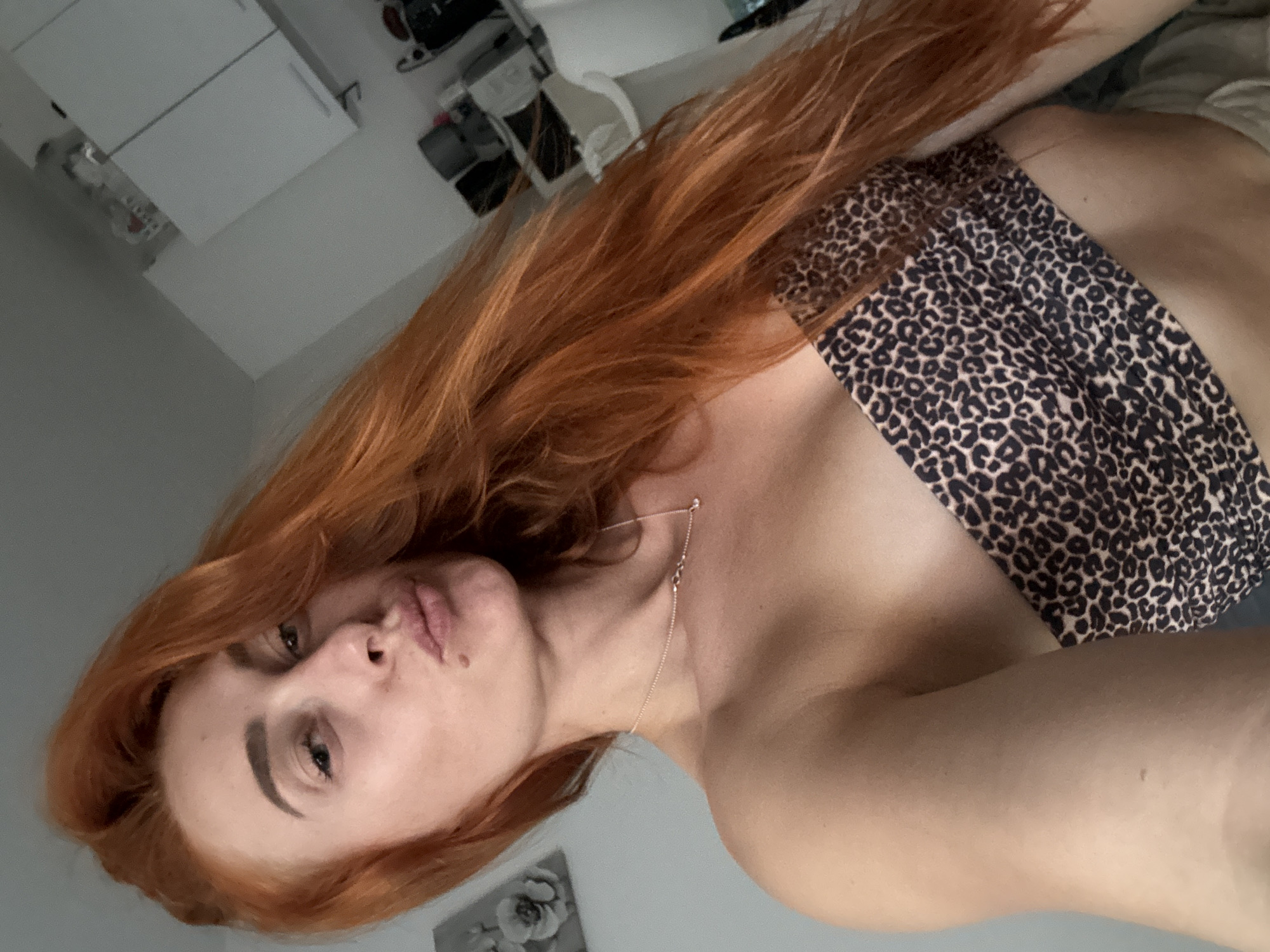 This media file is a close-up, vertically oriented selfie of a young woman with long, reddish-orange hair. She is wearing a leopard-print top or bandeau. Her face is turned towards the camera, with her lips pursed in a kiss. She is wearing a delicate necklace. The background appears to be a bedroom with a bed and some furniture, suggesting an indoor setting. The lighting is soft and diffused, indicating it might be daytime with indirect sunlight or artificial lighting. No visible text is present in the image. The overall mood conveyed is casual and intimate. Given the provided location context of Budva, Montenegro, it is possible this image was taken there.
