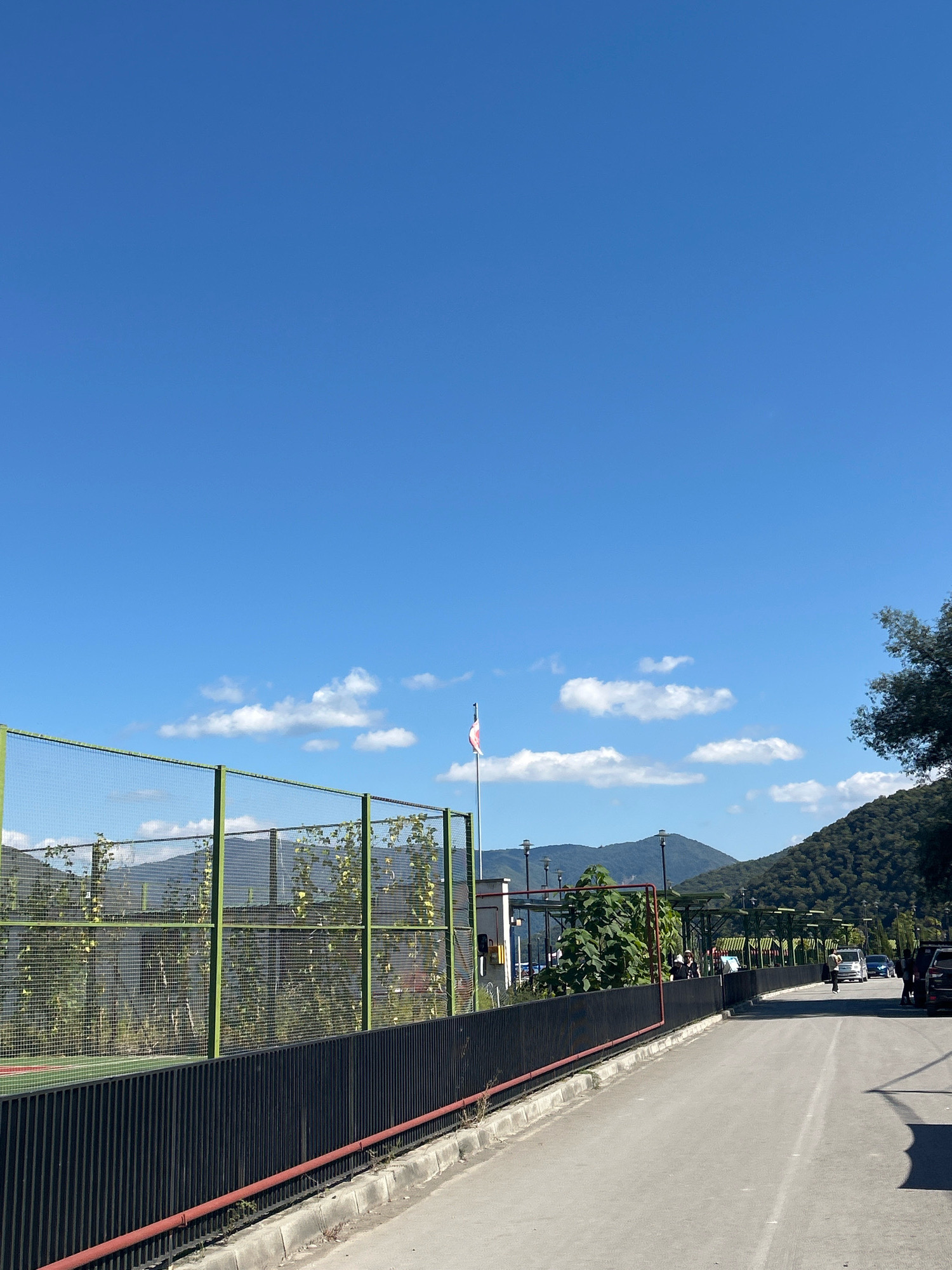 The image depicts an outdoor scene with a clear blue sky and scattered white clouds. In the foreground, a paved road runs from right to left, bordered by a black metal fence. Beyond the fence, a green-framed mesh fence encloses a sports court or similar area, with some vegetation visible behind it. A flagpole with a red and white flag stands near the mesh fence. In the background, there are trees, hills, and mountains. Several vehicles and a few people are visible on the road in the middle ground. The overall impression is of a sunny day in a semi-urban or recreational area. The provided context suggests the location is Shalikiantkari, Georgia, though this cannot be visually confirmed from the image alone.