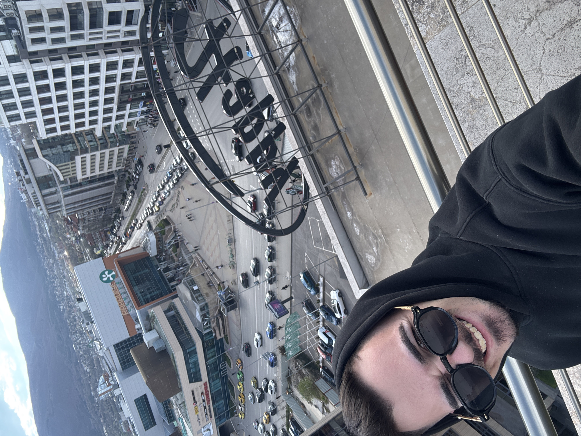 A selfie captures a man wearing sunglasses and a black hoodie, standing on a balcony with a metal railing. Behind him, the urban landscape of Sofia, Bulgaria, unfolds. The "Paradise Center" shopping mall is visible, along with the distinctive "Paradise" sign. The surrounding area features a mix of buildings, streets with vehicular traffic, and a backdrop of mountains in the distance. The shot is taken from a high vantage point, offering a panoramic view of the city.