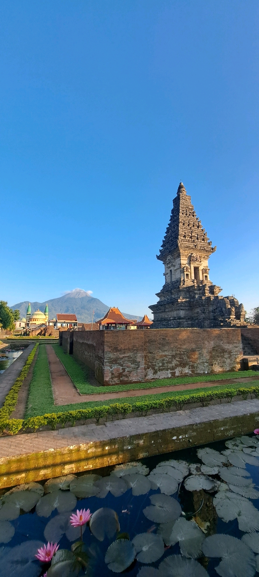In Prigen, Indonesia, a serene scene unfolds at sunrise. In the foreground, a tranquil pond teems with lily pads and two vibrant pink lotuses. Beyond the pond, a neatly manicured lawn borders a pathway that leads towards a weathered brick wall. In the mid-ground, the wall encloses a historical structure, a tiered stone pagoda. Behind the pagoda, a village is visible with traditional Indonesian architecture, including buildings with terracotta roofs and two minarets. In the distance, a majestic mountain, possibly Mount Arjuno, stands tall, its peak partially shrouded in a wispy cloud. The clear blue sky suggests a bright, clear day.