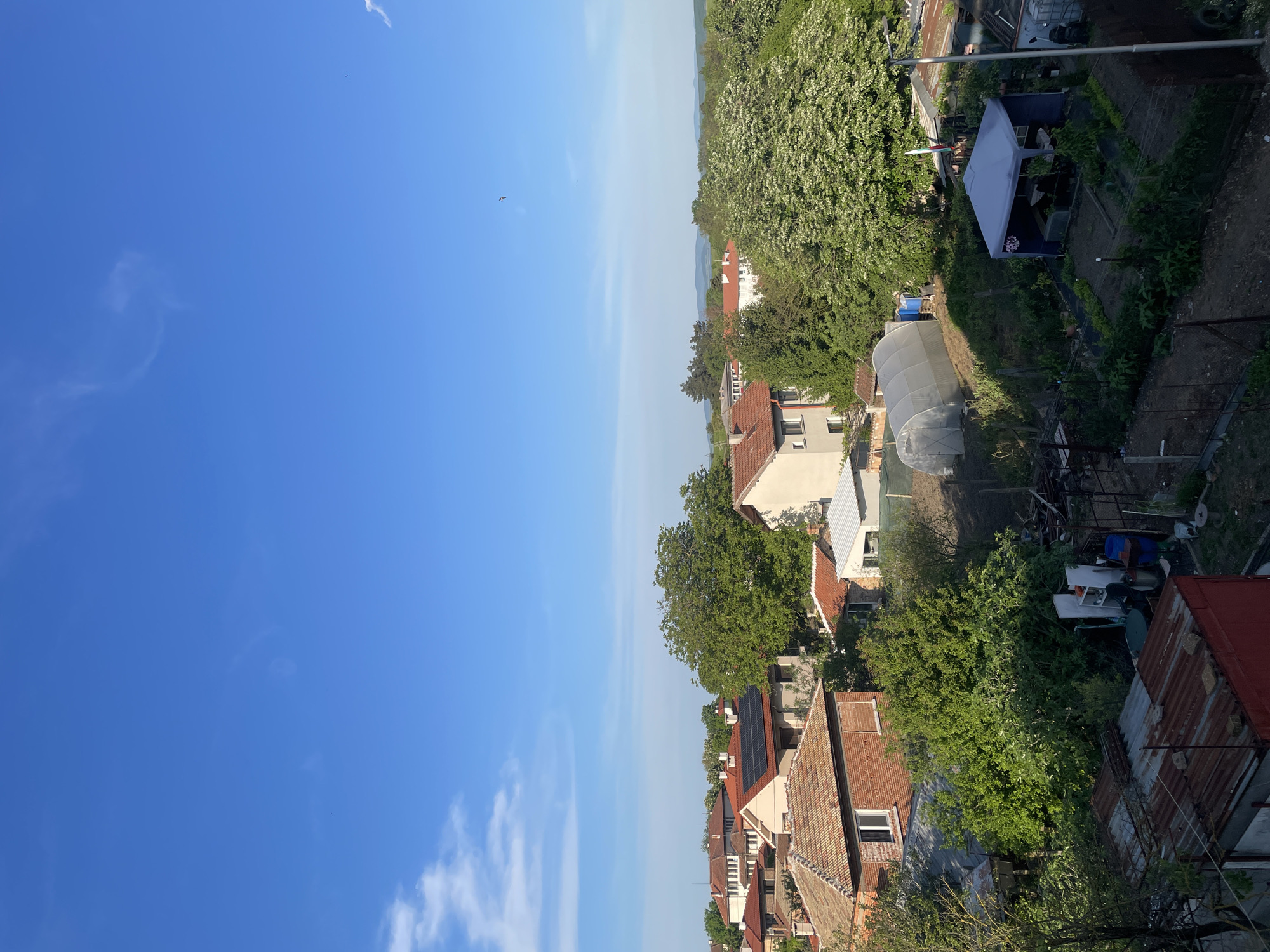 The image shows an elevated view of a residential area in Debelt, Bulgaria. The sky, predominantly blue with scattered clouds, takes up roughly half the frame. On the right, the scene is filled with houses, trees, and gardens. House rooftops are varied, including tile and corrugated metal. Some houses have solar panels. A greenhouse is also visible. There are no people visible. The overall environment is residential, possibly suburban.