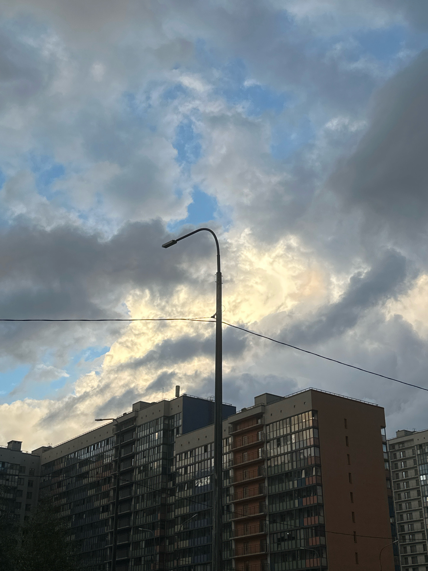 The image shows a modern apartment building against a cloudy sky with patches of sunlight breaking through. A street lamp stands prominently in the foreground, its dark pole contrasting with the bright sky. The building features numerous windows, reflecting the surrounding environment and the dramatic cloud formations. Power lines are strung horizontally across the frame, bisecting the view between the sky and the building. The scene is set in an urban environment, likely during the day, given the natural light.