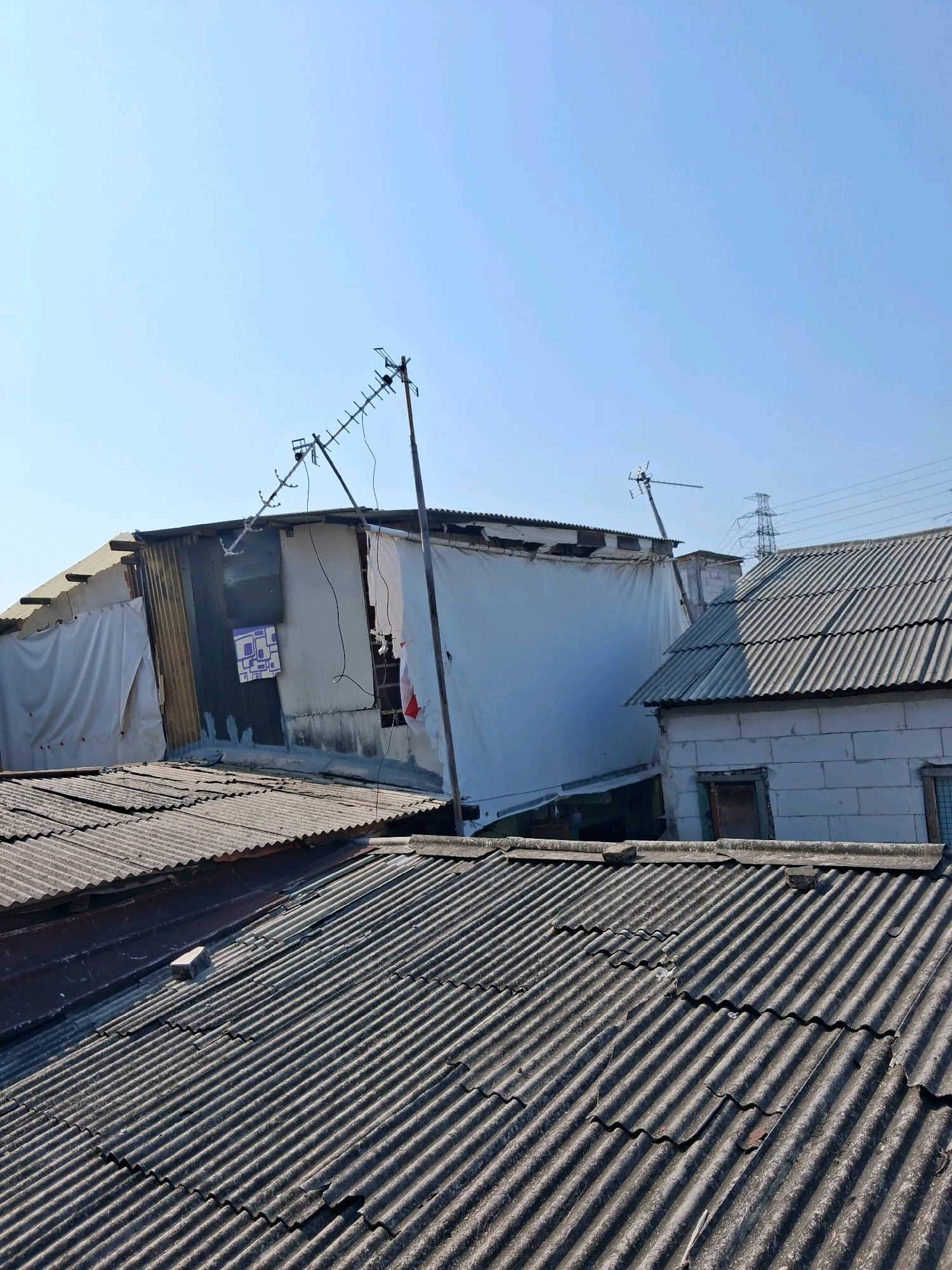 The image shows a view of rooftops in a densely populated area of North Jakarta, Indonesia, characterized by a mix of corrugated metal and possibly asbestos roofing materials, some of which appear aged and damaged. The architecture suggests informal settlements or older industrial areas. In the foreground, several layers of corrugated roofs dominate the view. In the mid-ground, a makeshift structure is visible, constructed with a combination of corrugated metal sheeting, white tarpaulin, and what appears to be concrete blocks. An old television antenna is mounted on a pole, leaning against this structure. A partially visible sign with purple geometric patterns is attached to the side of the building. The background reveals a power pylon and transmission lines, common infrastructure elements in urban environments. The sky is clear and blue, indicating sunny weather. The overall scene depicts a common urban landscape in parts of North Jakarta, where resourcefulness and makeshift construction are evident in response to housing needs.