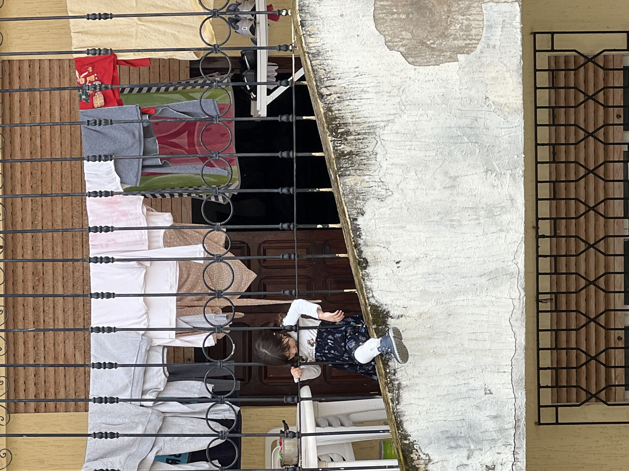 An outdoor scene depicts a child standing on a concrete ledge in the city of M'Diq, Morocco. The ledge is adjacent to a building with an exterior wall painted white. A black wrought-iron security grill covers a window to the right of the child. To the left of the child is a black wrought-iron railing with clothes hanging to dry. The clothes include items in red, grey, pink, white, and brown. A dark brown wooden door and furniture are visible behind the railing and clothesline. The child is wearing a long-sleeved white shirt, a dark blue patterned dress, white socks, and grey sneakers. The child has brown hair and appears to be looking down.