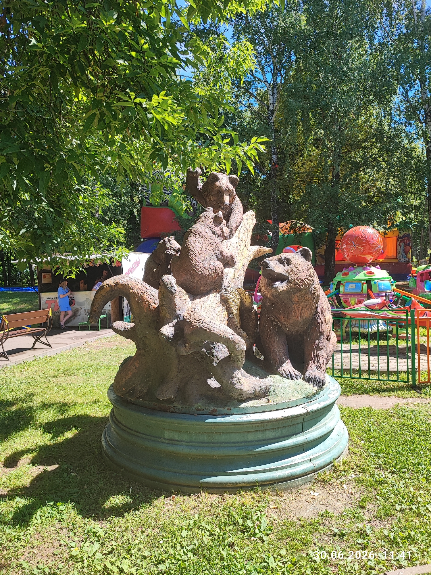 The image depicts a sculpture of bears in an outdoor setting. The sculpture, situated on a green circular base, features several bear figures arranged on what appears to be a stylized tree trunk. The scene is set in a park-like environment with lush green grass, trees, and sunlight casting shadows. In the background, elements of an amusement park are visible, including colorful rides and structures. To the left, there is a small kiosk or stall with people interacting and a park bench. The overall impression is that of a public recreational area, possibly within an amusement park or a park with children's attractions in the city of Lytkarino, Russia, as indicated by the timestamp in the bottom right corner which reads "30.06.2026 11:41" (though the year may be a misprint and likely refers to 2024 or another recent year, given the context of current date).
