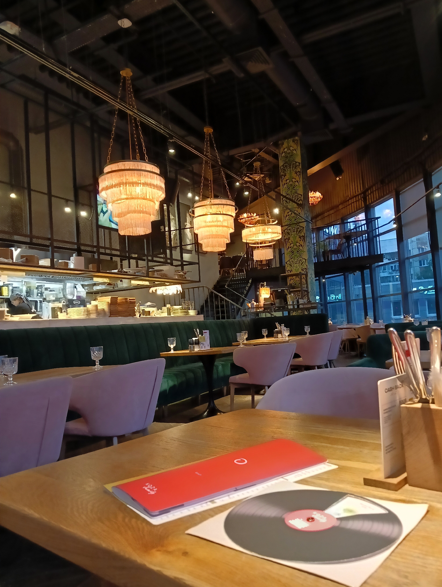The image shows the interior of a modern restaurant in Saint Petersburg, Russia. The scene is set at a wooden table in the foreground, where a red menu and a vinyl record are placed. In the background, plush green banquettes line the walls, with elegant pink chairs arranged around tables. The restaurant features a high ceiling with exposed industrial beams and ornate chandeliers. A bustling open kitchen is visible on the left, with chefs working behind a counter. Large windows on the right offer a view of the city at dusk, with buildings and streetlights illuminated. The overall ambiance suggests a sophisticated dining establishment.