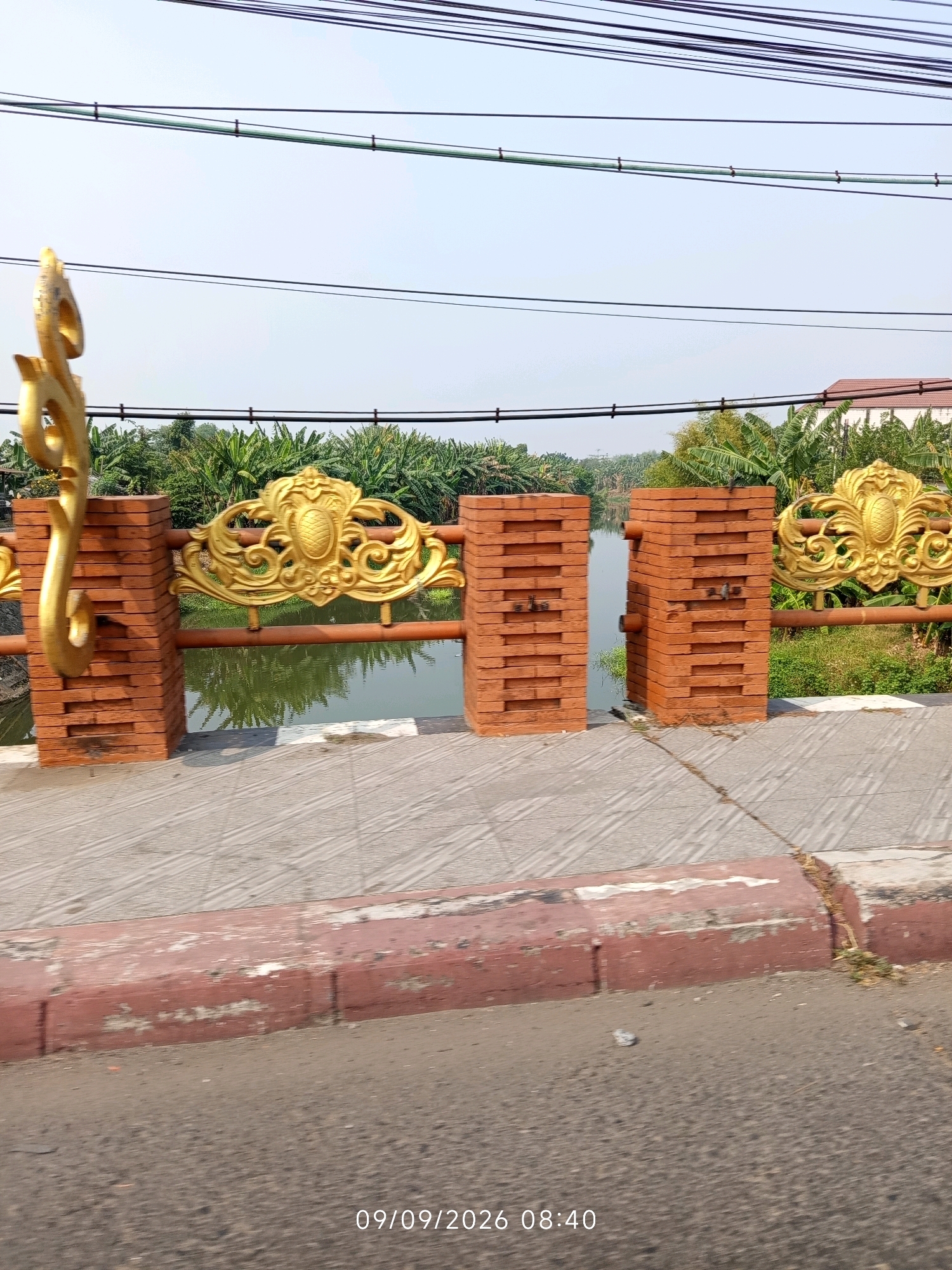 The image depicts a bridge railing with ornamental gold carvings and brick pillars, overlooking a body of water with lush green vegetation on the opposite bank. The railing appears to be part of a pedestrian walkway or bridge. In the foreground, a sidewalk with a red curb is visible, with asphalt road beyond it. Overhead power lines stretch across the frame. The scene is illuminated by natural daylight, suggesting it was taken outdoors. A timestamp in the lower right corner indicates "09/09/2026 08:40". The specific location in Mojokerto, Indonesia, is not explicitly identifiable from the image's content alone, but the overall setting suggests a possibly urban or semi-urban environment with a waterway.