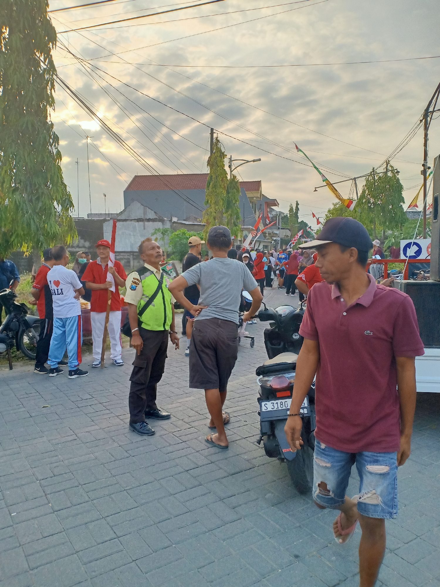 The image depicts an outdoor scene in the city of Sumber Tlaseh, Indonesia, likely during daytime given the lighting conditions. A group of people are gathered on a paved street, which is bordered by trees and buildings. In the foreground, a man wearing a maroon polo shirt and ripped denim shorts is walking away from the camera. To his left, a motorcycle is parked on the street.
Further into the scene, several individuals are interacting. One man, wearing a reflective vest over a light-colored shirt and dark pants, appears to be a security guard or official. He is looking upwards. Another man in a red shirt is holding a long pole with an Indonesian flag attached. Other people are dressed in casual attire, with some wearing white t-shirts and blue pants. Decorative flags and banners are visible in the background, suggesting a possible event or celebration. The sky is partly cloudy, with sunlight filtering through. The overall atmosphere appears to be casual and communal.