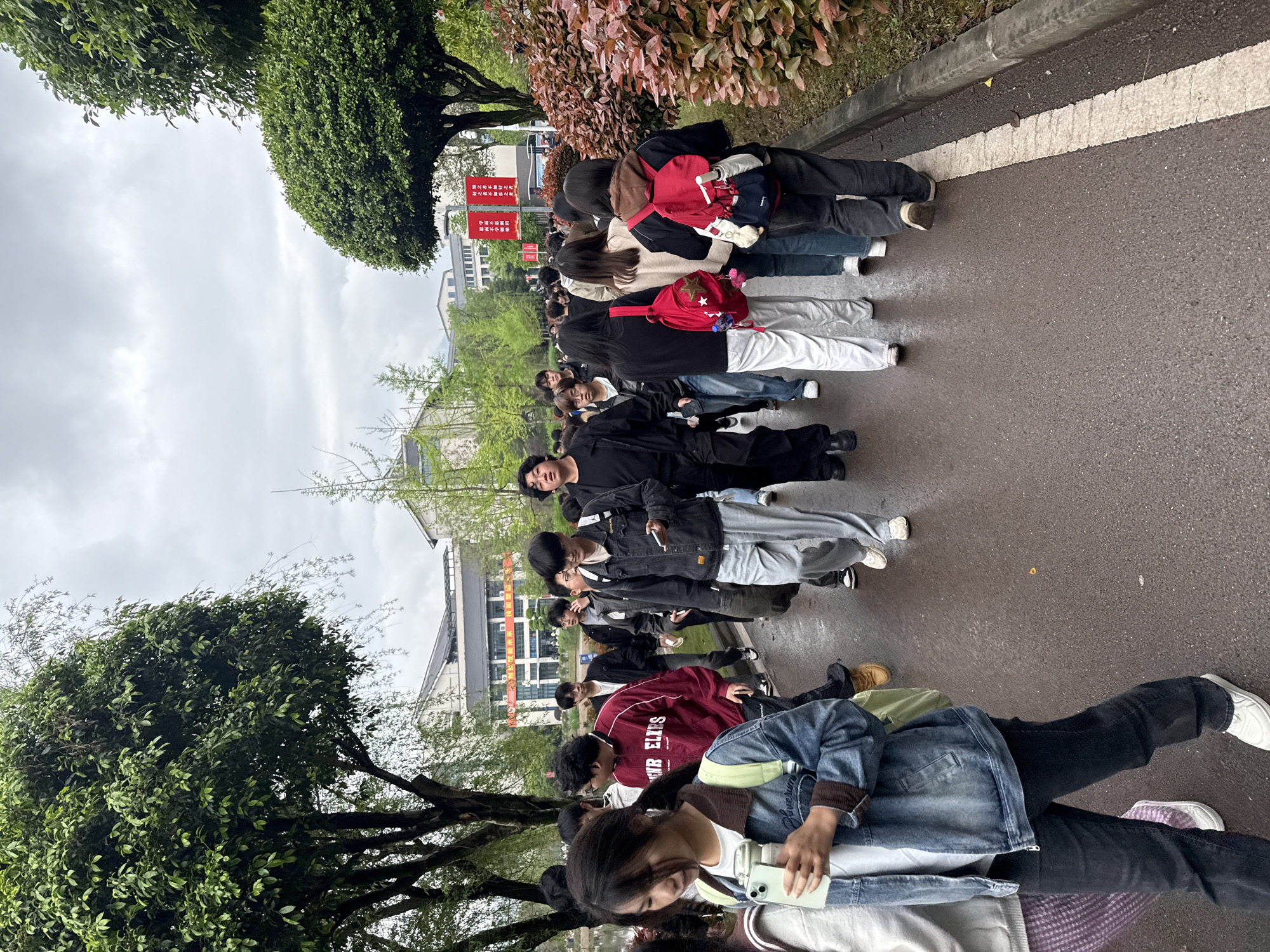 A medium eye-level shot depicts a large group of young people standing in a line on a dark, wet pavement, presumably in Jiangjin, China. The group extends from the bottom to the middle of the frame and then bends at an angle to the right. On the lower right side, a girl is holding a smartphone, looking down. The group is standing between trees, buildings, and signs, partially visible in the background.