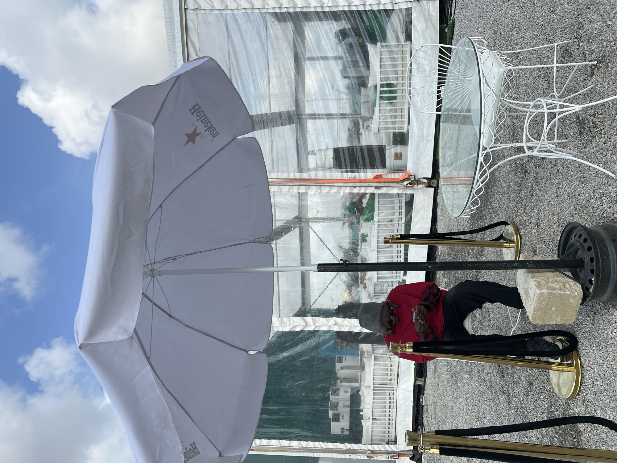 A man in a red shirt, black pants, a grey hat, and sunglasses is seated on a white stool behind a stanchion with a gold-colored base and a black rope. He is holding a mobile phone in his hands. A white Heineken umbrella provides shade. A clear, tent-like structure is visible behind him, and a white, wrought-iron table with a glass top stands nearby. The ground is covered in small grey stones. The location is likely an outdoor event space in Lagos, Nigeria.