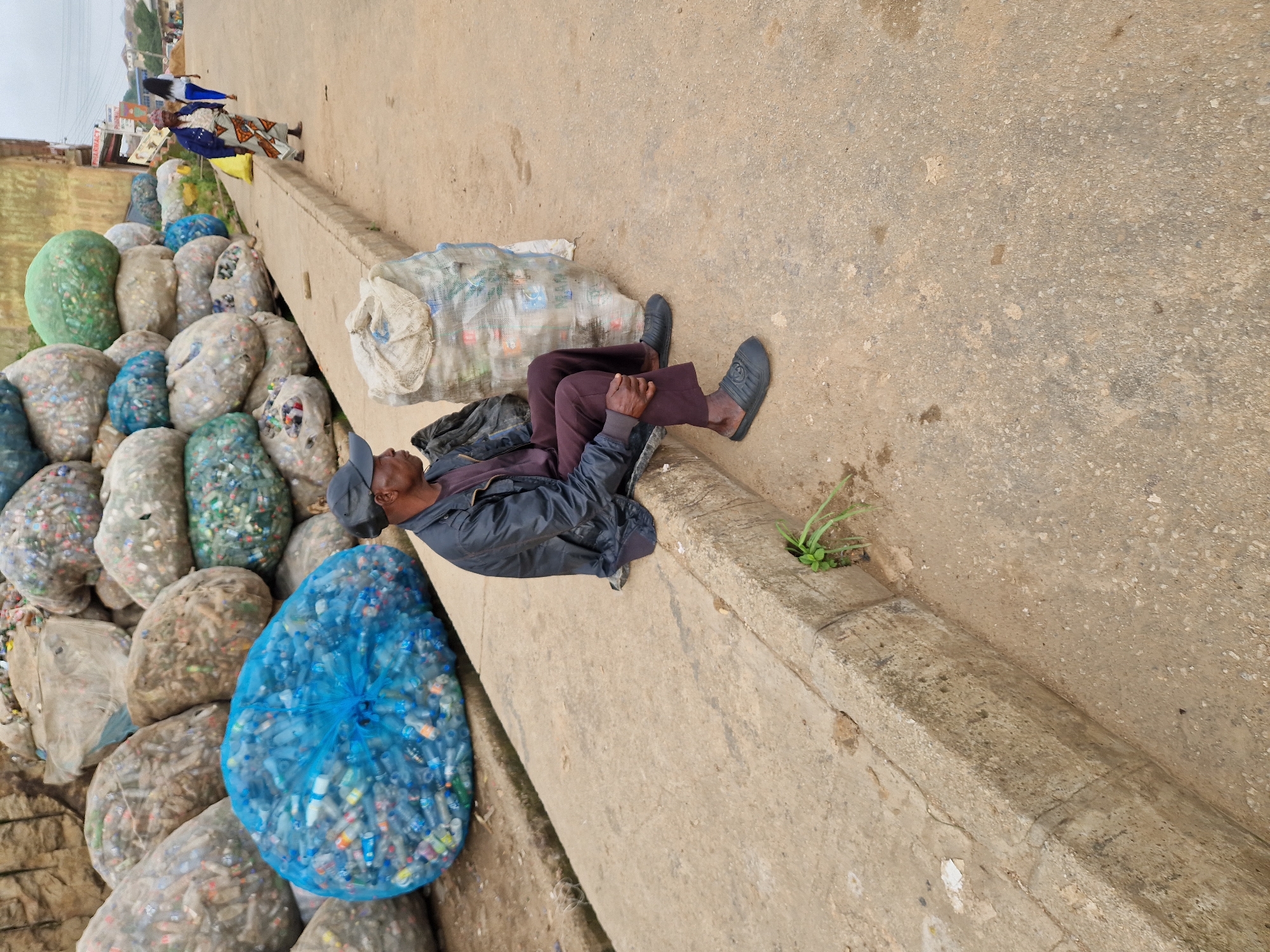 The image depicts a street scene in Kabong, Nigeria, likely a recycling or waste collection area. In the foreground, a man wearing a dark cap, a dark jacket, and maroon pants sits on a concrete curb. He is wearing black slip-on sandals. Next to him are two large, bagged bundles of what appear to be plastic bottles. Behind the man and to his left, a large blue mesh bag is overflowing with plastic bottles. Further in the background, more large, loosely packed bags of plastic bottles are piled high, creating a substantial mound. The environment is dusty and appears to be an outdoor setting with a building visible in the far background on the right, and a few other figures walking in the distance. The overall impression is one of informal waste management and recycling activities.