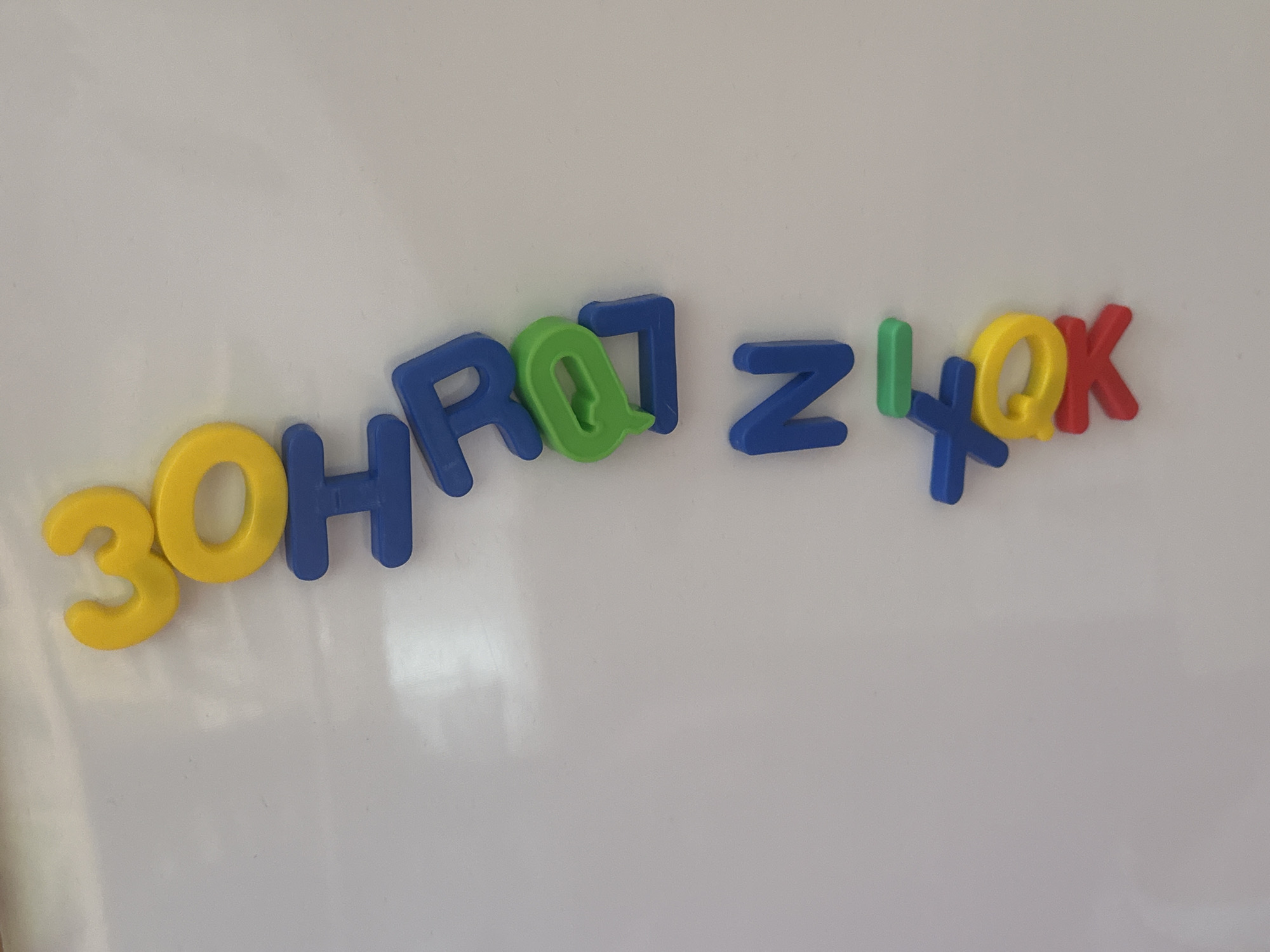 The image displays a collection of colorful magnetic letters and numbers on a white surface, likely a whiteboard or a similar material. The items are arranged in a scattered, somewhat haphazard manner across the frame.
From left to right, the visible characters are: a yellow number '3', followed by a yellow letter 'O', a blue letter 'H', a blue letter 'R', a green letter 'Q', a blue letter 'J', a blue letter 'Z', a green vertical bar or possibly a lowercase 'l', a blue letter 'X', a yellow letter 'Q', and a red letter 'K'. The letters and numbers appear to be made of plastic and have a matte finish. The white background is smooth and shows some subtle reflections and shadows, suggesting it is a flat, illuminated surface. There are no people or other objects present in the image. The overall scene is static and devoid of any activity.