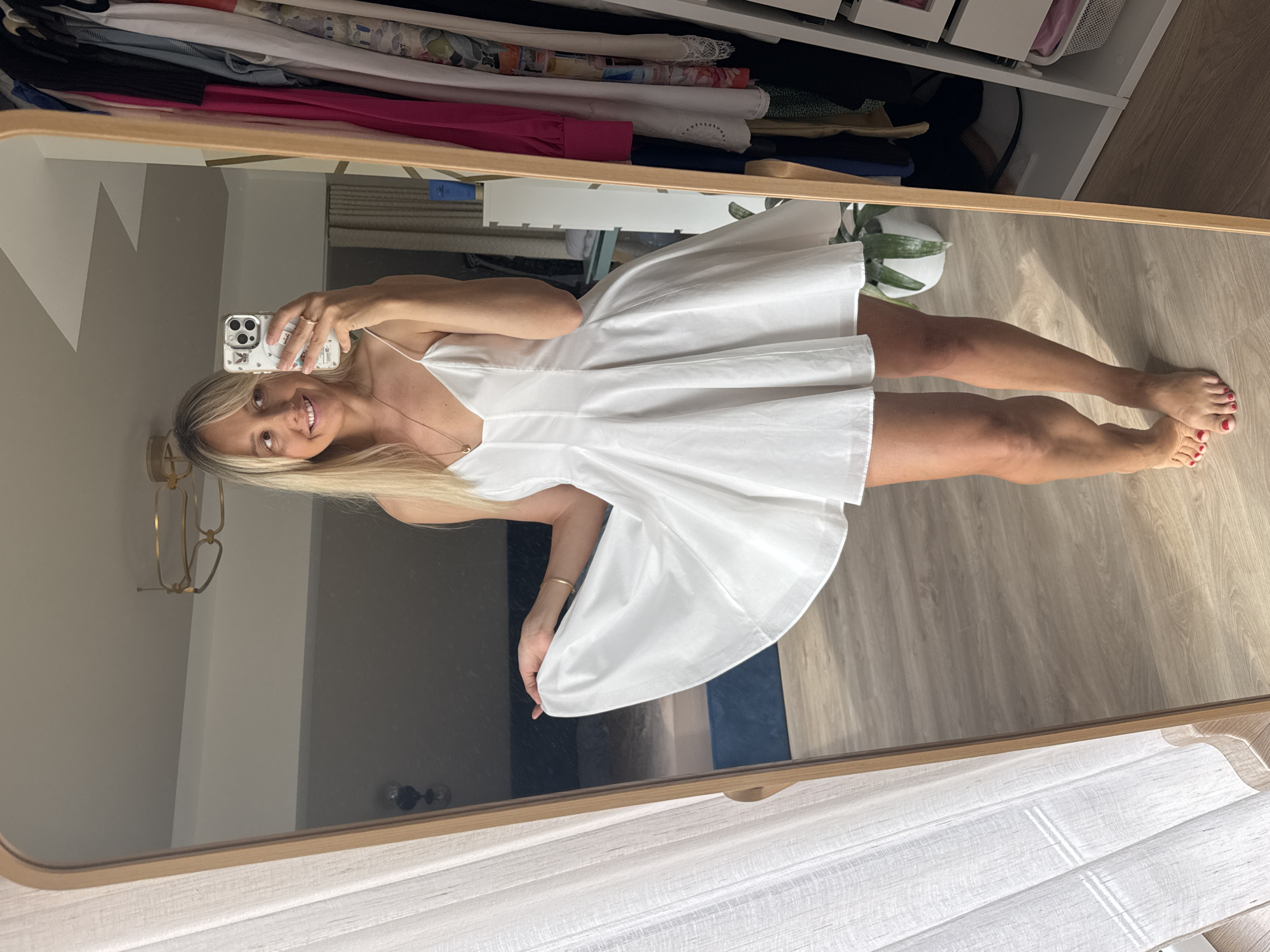 A blonde woman stands in front of a full-length mirror, taking a selfie with her phone. She is wearing a white slip dress with thin straps and a flared skirt. The mirror reflects her upper body and the room behind her, which appears to be a bedroom with a bed and curtains. To the right of the mirror, a white closet filled with clothes is visible. The woman is smiling and holding her phone up to capture the image. The floor is made of wooden planks. The location is not identifiable as Carnikava, Latvia.