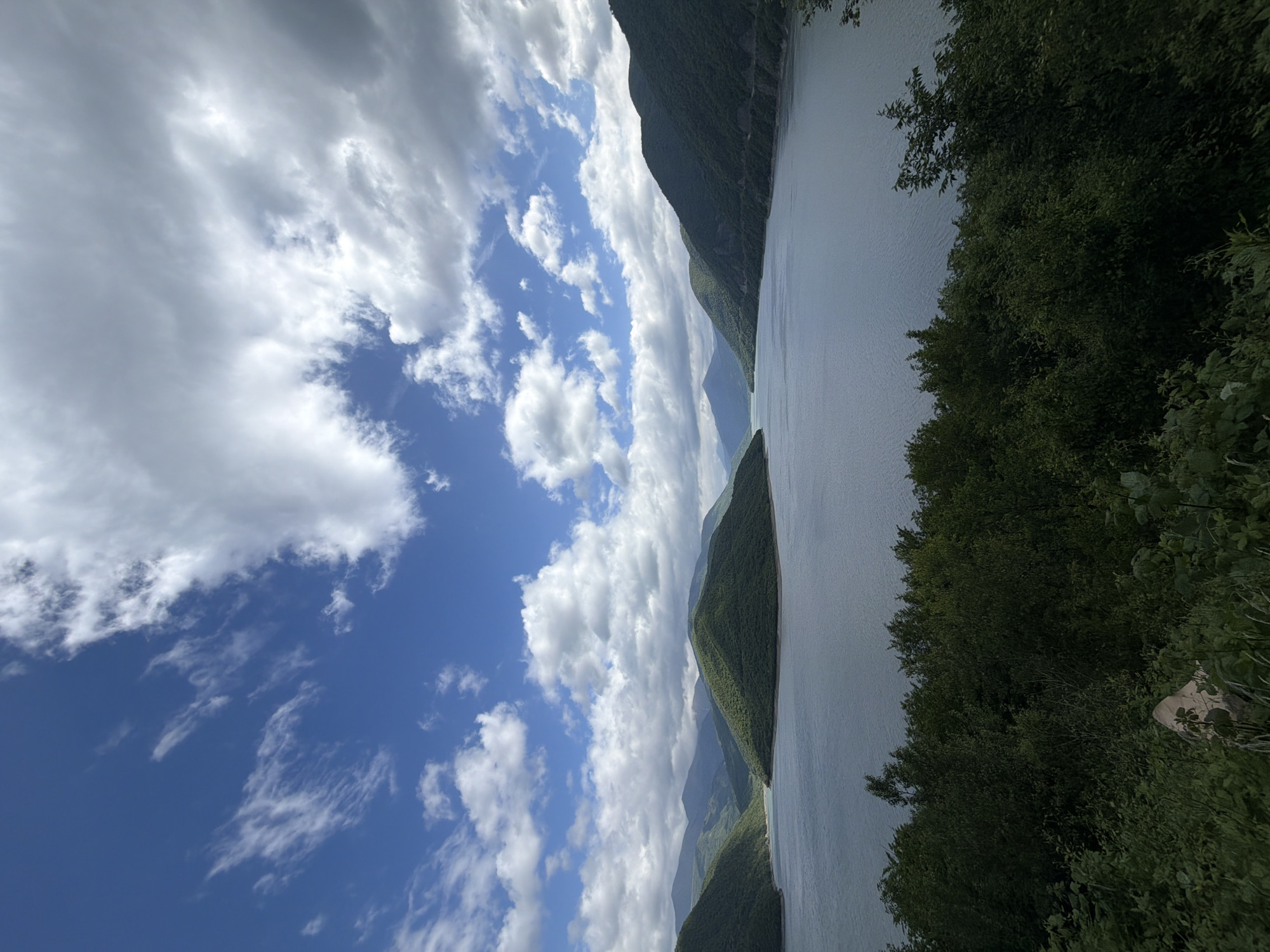 The image depicts a body of water surrounded by mountainous terrain. The sky is blue with scattered white clouds. Dense green foliage is present along the right edge of the frame. The location is Aranisi, Georgia. The mountains are tall and covered in greenery. The water surface is smooth and reflective. No people or animals are visible.