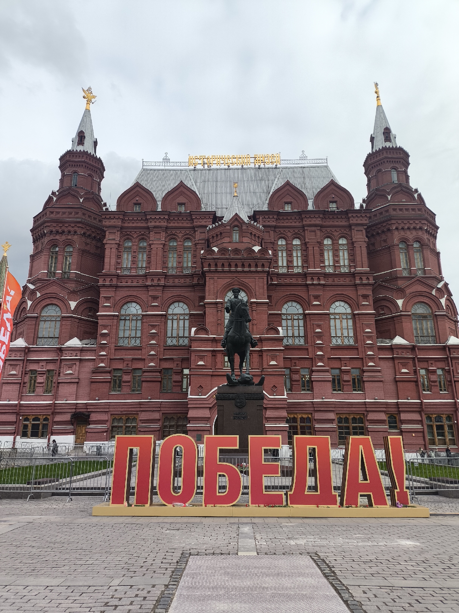 An outdoor, eye-level shot captures the State Historical Museum on Red Square in Moscow, Russia, under an overcast sky. The majestic building, constructed from red brick, features a Neo-Russian architectural style with intricate facades, numerous windows, and prominent spires topped with golden double-headed eagles. Golden letters spelling "ИСТОРИЧЕСКИЙ МУЗЕЙ" (Historical Museum) are visible above the central entrance.
In the foreground, a large, freestanding sign proudly displays the word "ПОБЕДА!" (Victory!) in prominent red letters with golden borders, resting on a golden base. Small red items, possibly flowers, are placed around the base of the letters, indicating a commemorative occasion like Victory Day (May 9th). Behind this sign, an equestrian statue, likely depicting Marshal Georgy Zhukov, stands on a dark pedestal.
The ground is paved with traditional cobblestones, and metal crowd barriers are set up across the square, suggesting preparation for or aftermath of a public event. A red banner, partially visible on the left, also bears the word "ПОБЕДА!". The gray, cloudy sky indicates daytime, though without direct sunlight. A few indistinct figures can be seen near the museum and behind the barriers in the distance. The overall scene conveys a sense of national pride and commemoration.