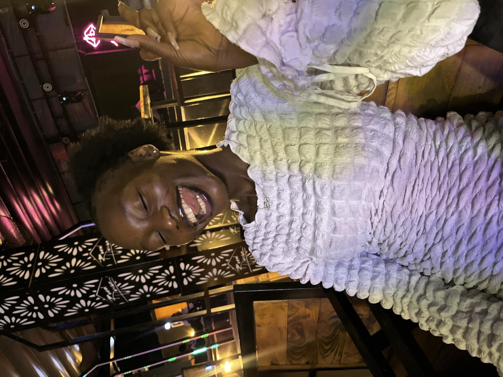 A young Black woman with her eyes closed and mouth wide open in laughter is pictured in a dimly lit indoor setting, likely a restaurant or bar in Howayi, Nigeria. She is wearing a textured white dress with puffed sleeves and a small silver necklace. Her right hand is raised, with fingernails painted and extended. Behind her, a decorative black metal panel with a floral cutout pattern is visible, illuminated by soft lights. In the background, neon signs and ceiling lights suggest a vibrant nightlife atmosphere. The woman's posture and expression convey a moment of genuine joy and amusement.