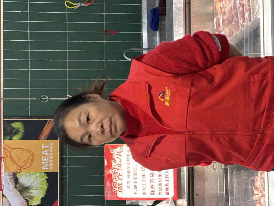 An adult female, appearing to be of East Asian descent and middle-aged, stands centered in the frame, facing the camera with a neutral expression. She wears a red turtleneck shirt under a red zippered vest adorned with a "家家欣" (Jia Jia Xin) logo on the left chest. The setting is identified as a market or supermarket in Jiangjin, China.
Behind her, a dark green tiled wall with white vertical grid lines is visible. To the upper left, a poster displays "MEAT" in English, accompanied by Chinese characters and illustrations of lettuce, chicken feet, and other food items. Below this, partially obscured by the woman, is a red sign with white Chinese text, including "温馨提示" (Warm Reminder) regarding fresh food.
To the right of the woman, various metal fixtures are present, including hanging hooks and a curved faucet above a sink or basin area. Yellow-handled scissors and a red tool hang on the green wall. Below the counter level, through a clear glass display, multiple trays of packaged raw meat products are visible. Two folded cloths, one blue and one brown, rest on a raised surface to the right.