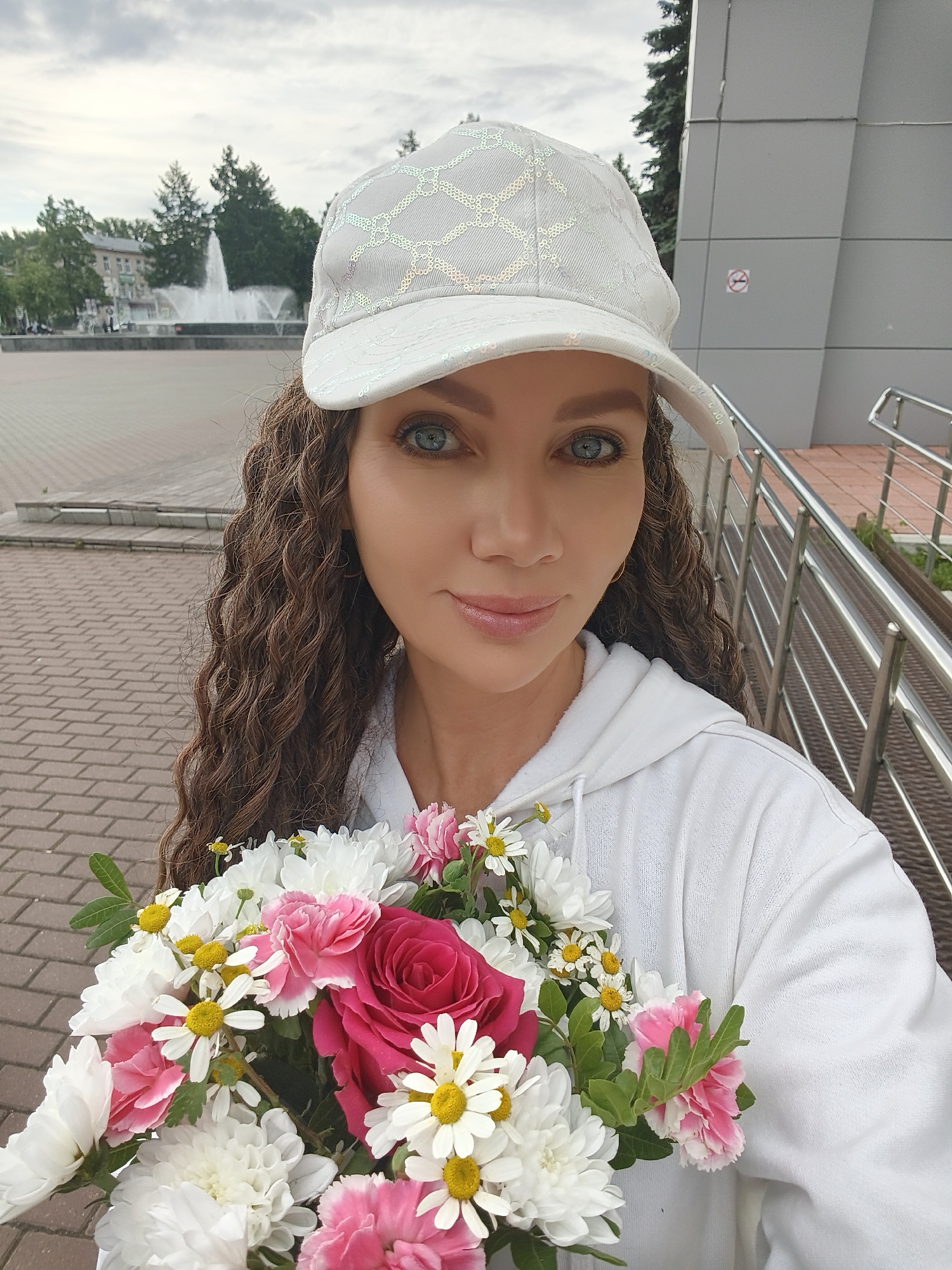 A woman with long, curly brown hair and blue eyes is holding a bouquet of flowers and wearing a white baseball cap and a white hooded sweatshirt. She is standing outside in front of a building with a gray, modern facade and a metal railing, possibly on a ramp or staircase. In the background, there is a fountain in a square, trees, and buildings. The scene appears to be in Chaikovsky, Russia, given the presence of a fountain and buildings characteristic of the city.