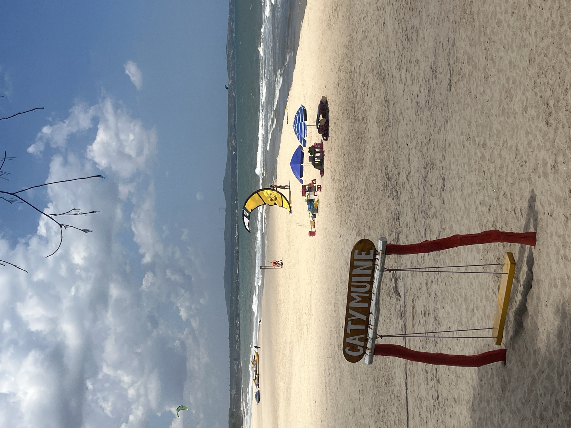 This is a photograph taken at a beach in Phu Thuy Ward, Vietnam. The image features a sandy beach with a gentle shoreline and waves crashing onto the shore. In the foreground, a wooden swing set with red supports and a yellow seat is visible, adorned with a rustic wooden sign that reads "CATYMUINE."
Scattered across the beach are several beach umbrellas, some striped blue and white, and others solid blue, providing shade. There are also a few beach chairs. In the distance, a kite surfer is visible against the backdrop of the ocean and a distant coastline with hills. Other kites can be seen in the sky and near the water. The sky is partly cloudy with some areas of bright blue, suggesting it is daytime with fair weather. The overall scene depicts a relaxed beach atmosphere, possibly a popular spot for water sports and recreation. The presence of the swing set and the sign suggests it might be a designated area or a resort.