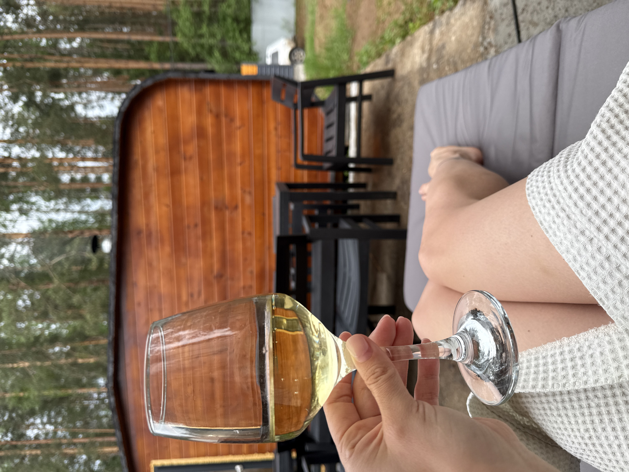 The image depicts a person holding a wine glass filled with a light-colored liquid, likely white wine. The individual is reclining on a grey cushioned lounge chair, wearing a white waffle-textured garment, with bare legs visible.
In the background, there is a wooden barrel-shaped sauna structure. Several black chairs are stacked behind the lounge chair. Trees are visible in the distance, suggesting a wooded or natural setting. The ground appears to be a concrete patio.
While the image is a photo, there is no way to tell the exact location, or the validity of it being in Chaykovsky, Russia.