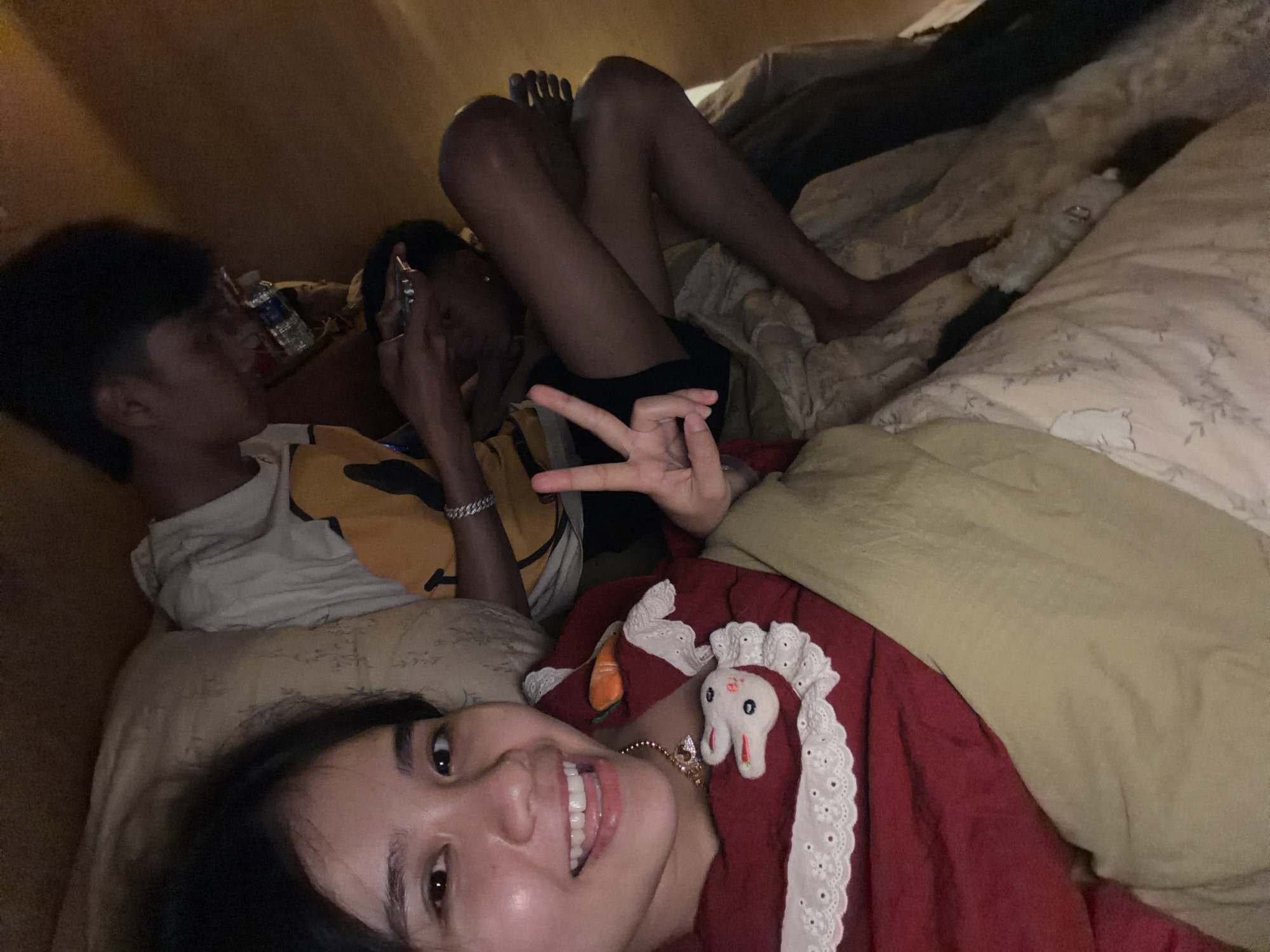 The image shows three young people relaxing indoors, likely in a bedroom setting, under dim, warm lighting that suggests evening or nighttime in Măng Đen, Vietnam.
In the foreground, a young woman lies on her back, looking directly at the camera with a cheerful, open-mouthed smile. She has dark hair and is wearing a red top adorned with white lace trim, featuring a small white bunny plushie and an orange carrot-shaped accessory. Her right hand is raised, making a peace sign towards the camera.
Behind her, to the left, another young person, appearing male, is seen lying on their side with their head turned away from the camera. They have dark, curly hair and are dressed in a white t-shirt layered over a yellow top. A plastic water bottle is visible near their head.
Further in the background, a third individual is partially visible, primarily their legs and an arm. This person appears to be lying down, wearing shorts, with one hand visible near the second person's head, possibly holding a phone.
The scene depicts a casual and relaxed gathering, with the individuals appearing comfortable and at ease. There is no visible text in the image.