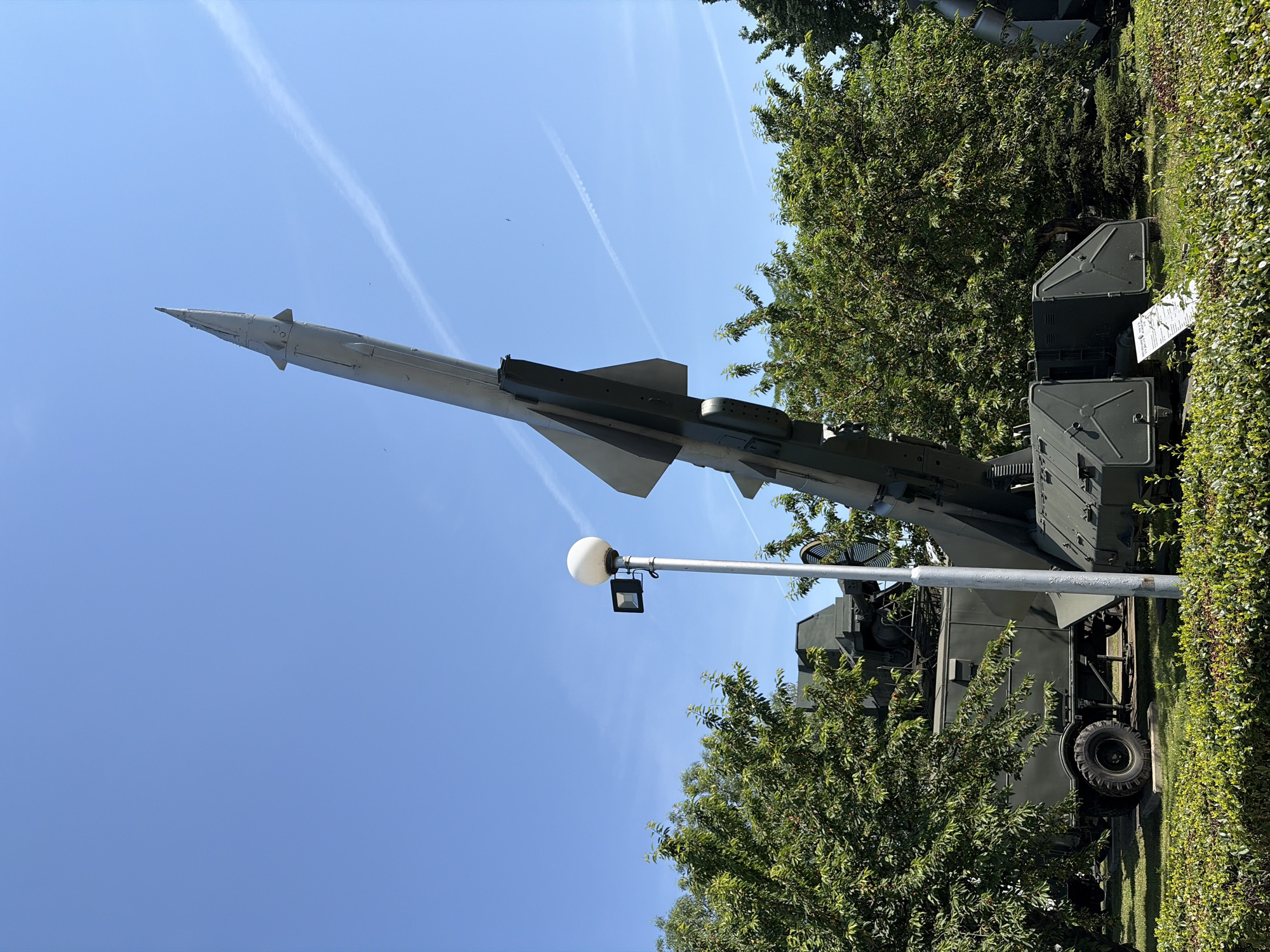 This is a photograph of a surface-to-air missile launcher, likely a Soviet-era SA-2 Guideline, displayed at the Museum of Aviation in Krumovo, near Plovdiv, Bulgaria. The missile is angled upwards towards a clear blue sky with faint contrails. The launcher is mounted on a green vehicle with large tires. In the foreground, there are trimmed green bushes. A streetlamp with a spherical light fixture and a smaller spotlight is positioned to the left of the missile. The overall environment appears to be an outdoor exhibition area with trees visible in the background.
