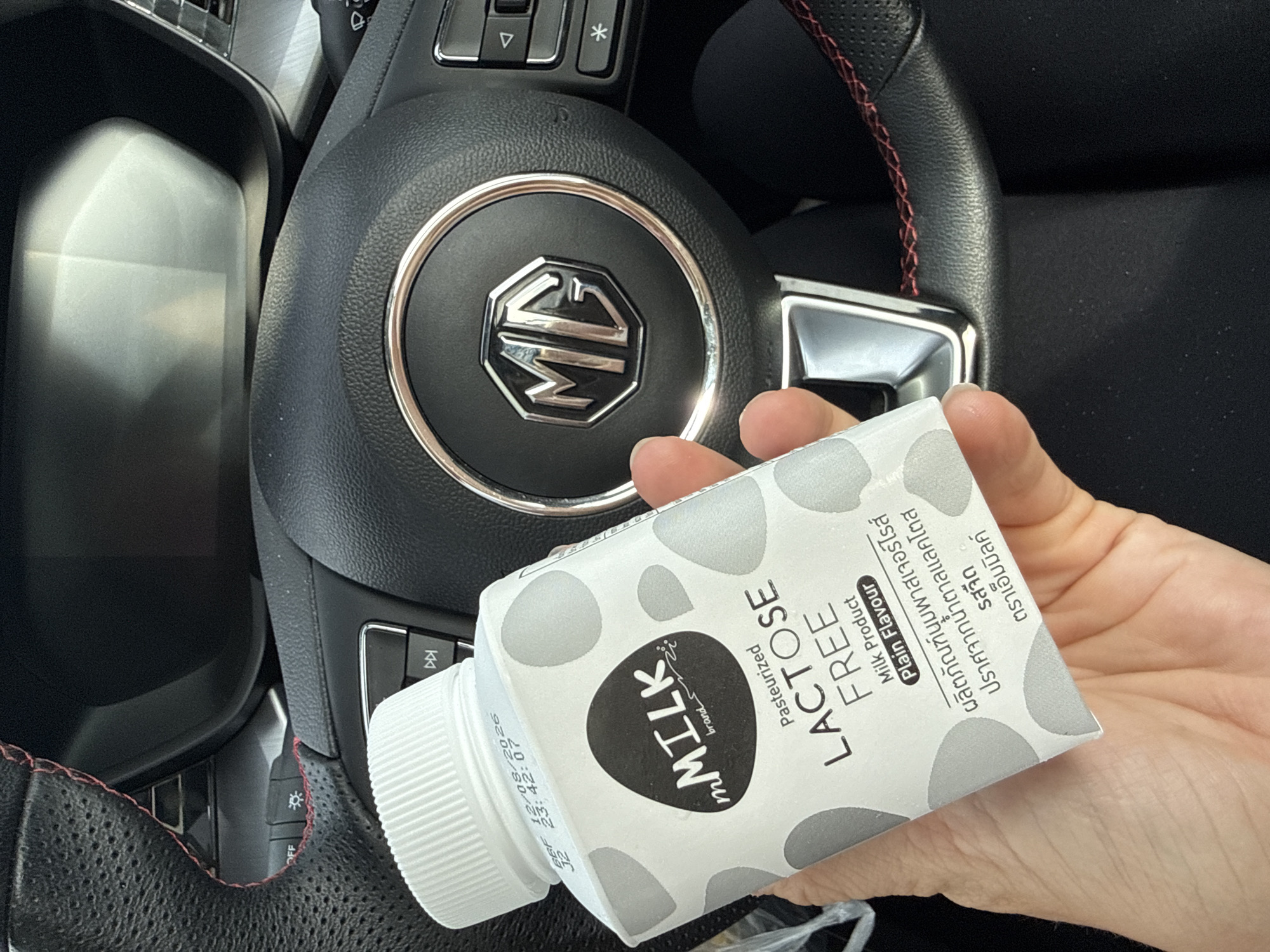 The image shows a hand holding a carton of mMILK brand Pasteurized Lactose Free milk. The carton has a gray and white polka dot pattern and text in English and Thai. The text in English reads "mMILK brand", "Pasteurized", "LACTOSE FREE", "Milk Product", and "Plain Flavour". The text in Thai, which is not fully legible, appears to describe the product. The background is out of focus but appears to be the interior of a car, with a steering wheel visible. The MG logo is visible on the steering wheel. The image is taken from a close-up perspective, focusing on the milk carton and the hand holding it. The lighting suggests it is daytime. Based on the visible text and common product availability, this image was likely taken in Ko Samui District, Thailand, where such products are commonly sold.