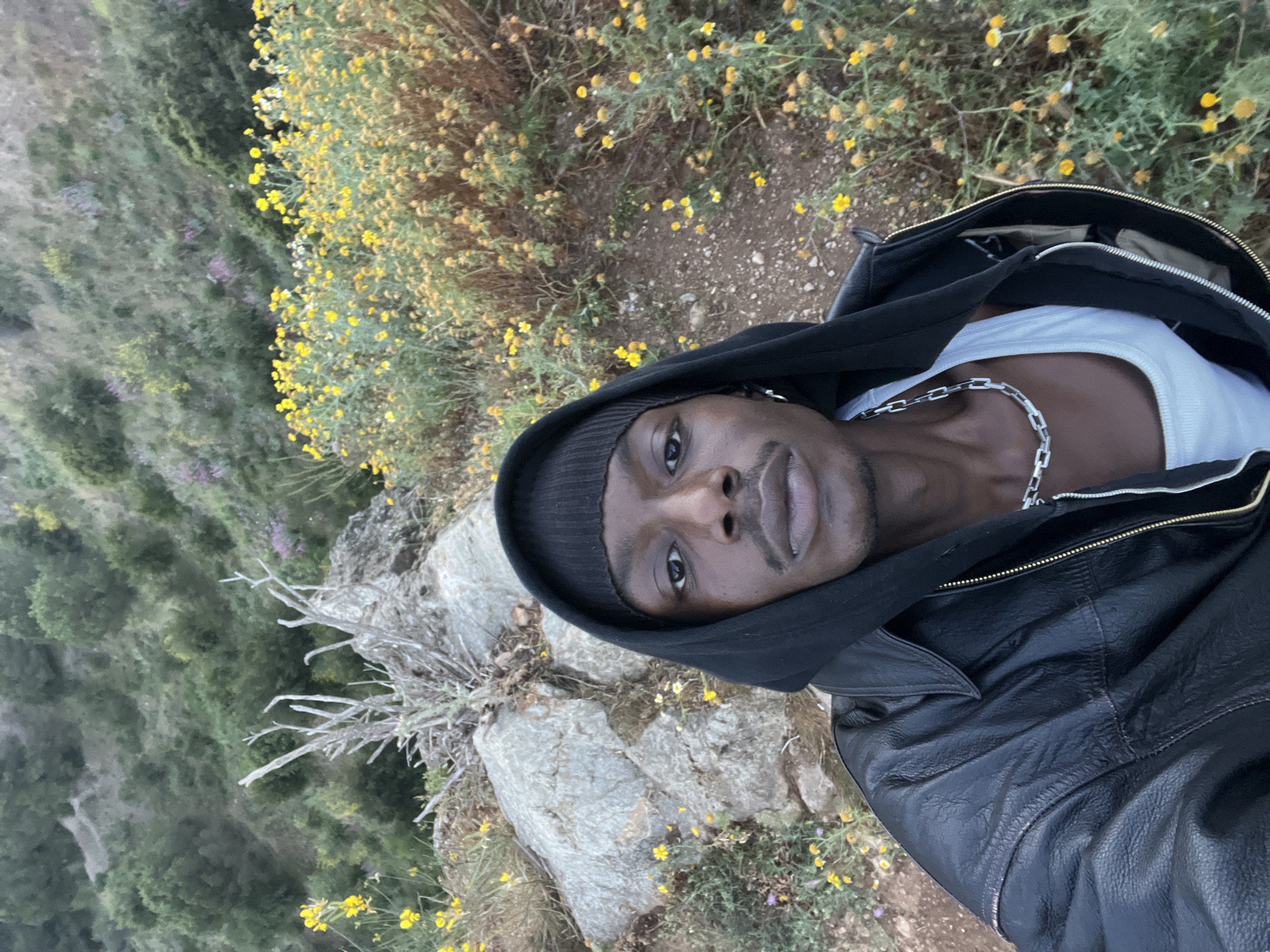 A medium-shot, self-portrait of a Black male wearing a black beanie, a black hooded leather jacket, and a white tank top with a silver chain. He appears to be standing on a rocky outcrop with yellow wildflowers. In the background is a verdant hillside. The photograph does not appear to be taken in Barcelona, Spain, as the environment does not match the urban setting of the city.