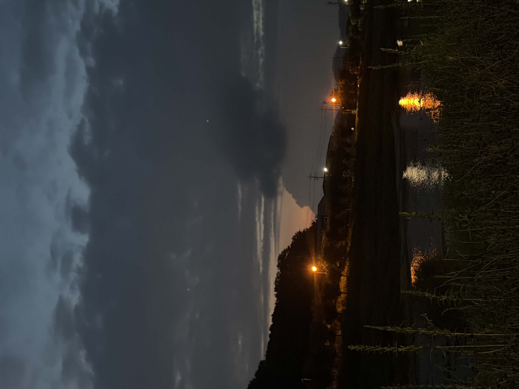 A nighttime landscape scene in Dongducheon-si, South Korea, shows a body of water in the foreground with reeds and grasses on the bank. The water reflects the orange glow of streetlights and the sky. In the middle ground, a tree-lined hill is silhouetted against a dusky sky with dark clouds. Several streetlights illuminate the area along the road at the base of the hill. The sky is dark and cloudy with a hint of twilight orange near the horizon. Two small, bright points of light are visible in the sky, possibly distant lights or celestial bodies.