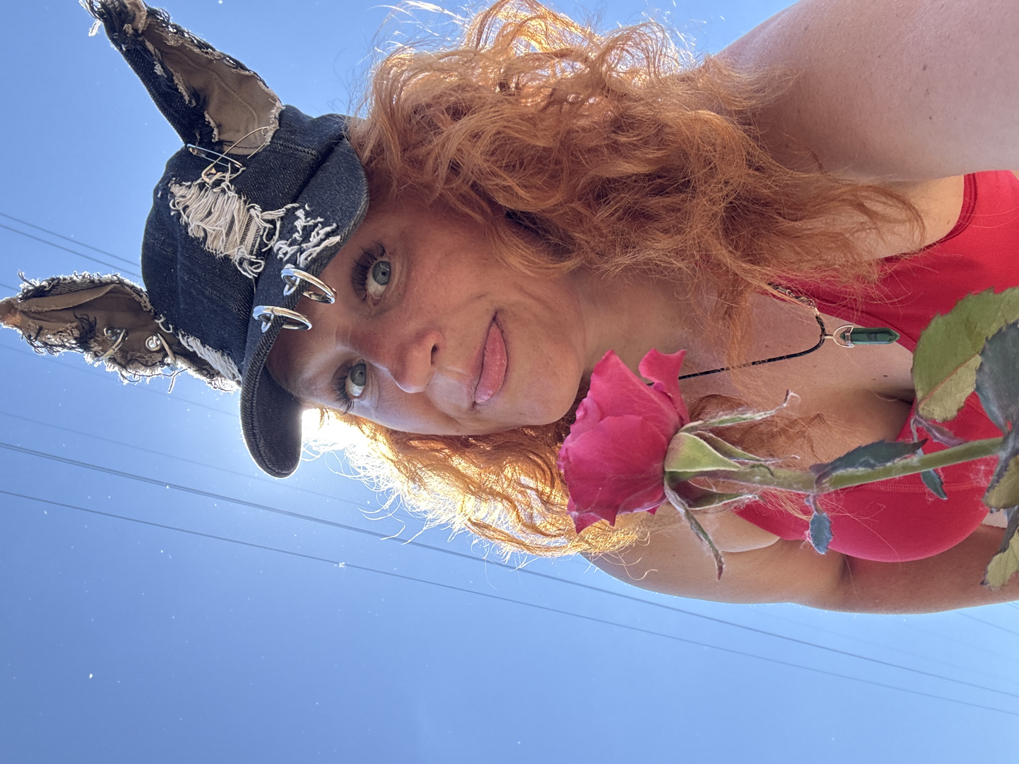A woman with vibrant red, curly hair and a custom denim hat with ragged edges and attached cat ears is captured in a close-up, outdoor selfie. The hat is adorned with silver piercings, including two heart-shaped hoops and safety pins. She is wearing a red top and a necklace with a green, crystalline pendant. A pink rose is held close to her face, partially obscuring her left cheek. Her right eye is wide open, and her tongue is playfully sticking out. The background reveals a clear blue sky with power lines stretching across it. The lighting suggests it is daytime with the sun shining brightly, creating a halo effect around her hair. The image appears to have been taken at a high angle, looking up at the subject. There is no visible indication of the location being in the city of Chaikovsky, Russia.
