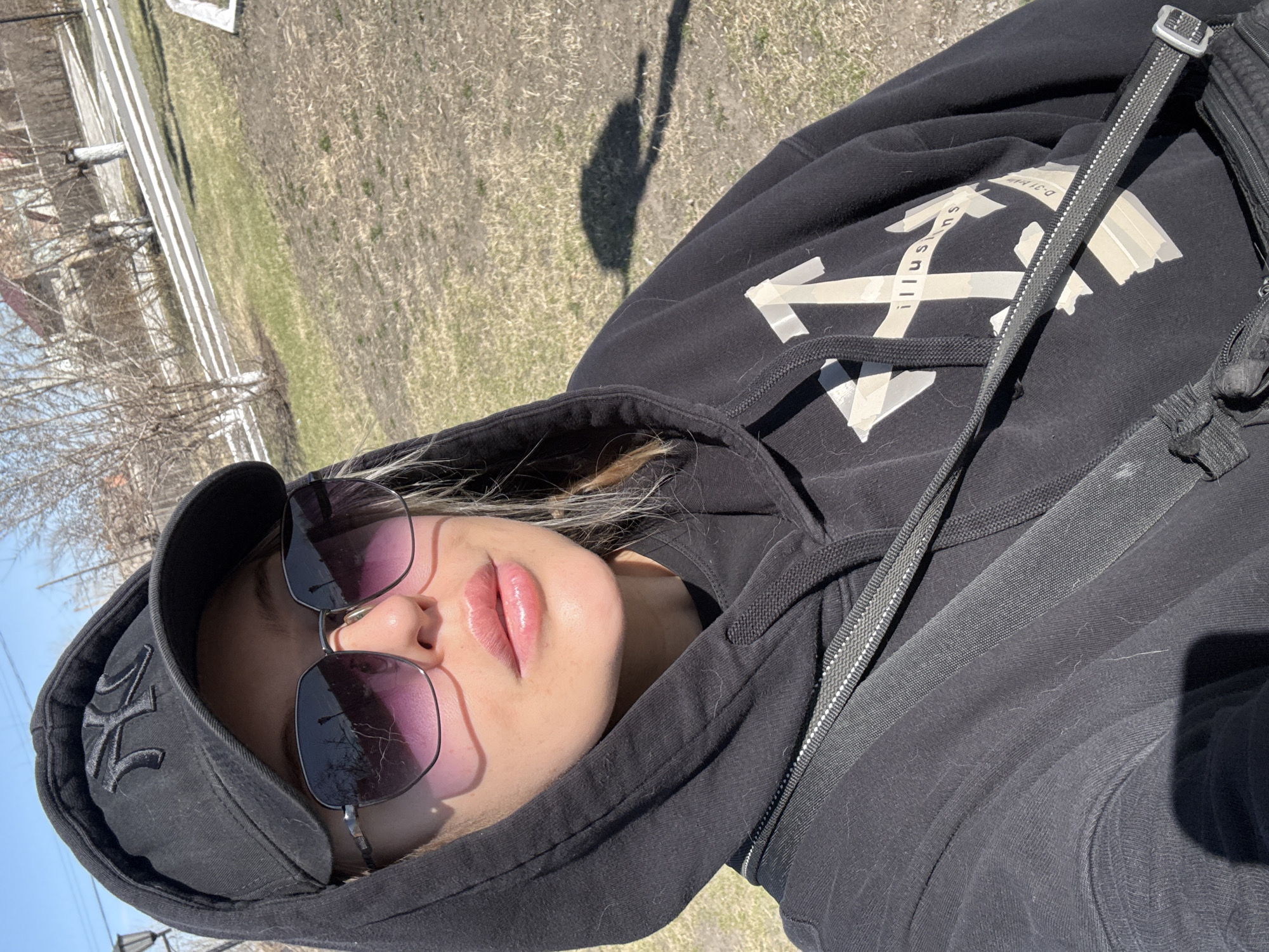 A young person wearing a black hoodie and a black baseball cap with the New York Yankees logo, is angled with their head towards the bottom left corner of the image. They also wear sunglasses with a dark tint. A black shoulder strap runs across the hoodie. The background shows a green grassy area, likely a park or field, with trees in the distance. A shadow cast by the person is visible on the ground. The specific location in the city of Borzya, Russia, cannot be determined from this image alone.