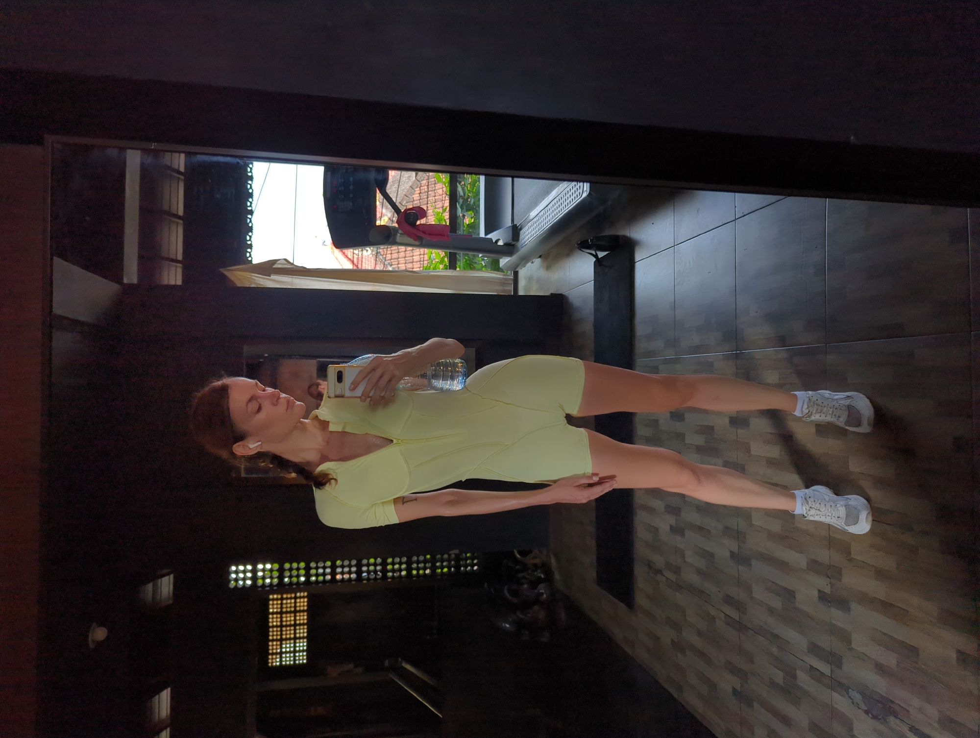 A woman in a light-yellow exercise outfit and white sneakers is reflected in a mirror inside what appears to be a workout space. She is holding a smartphone in her right hand, taking a selfie. Next to her, slightly out of focus, is a clear water bottle. The space appears to be indoors, with wooden floors and a treadmill visible behind her. Outside, reflected in the mirror, is greenery and a building. The location is Mengwi, Indonesia.
