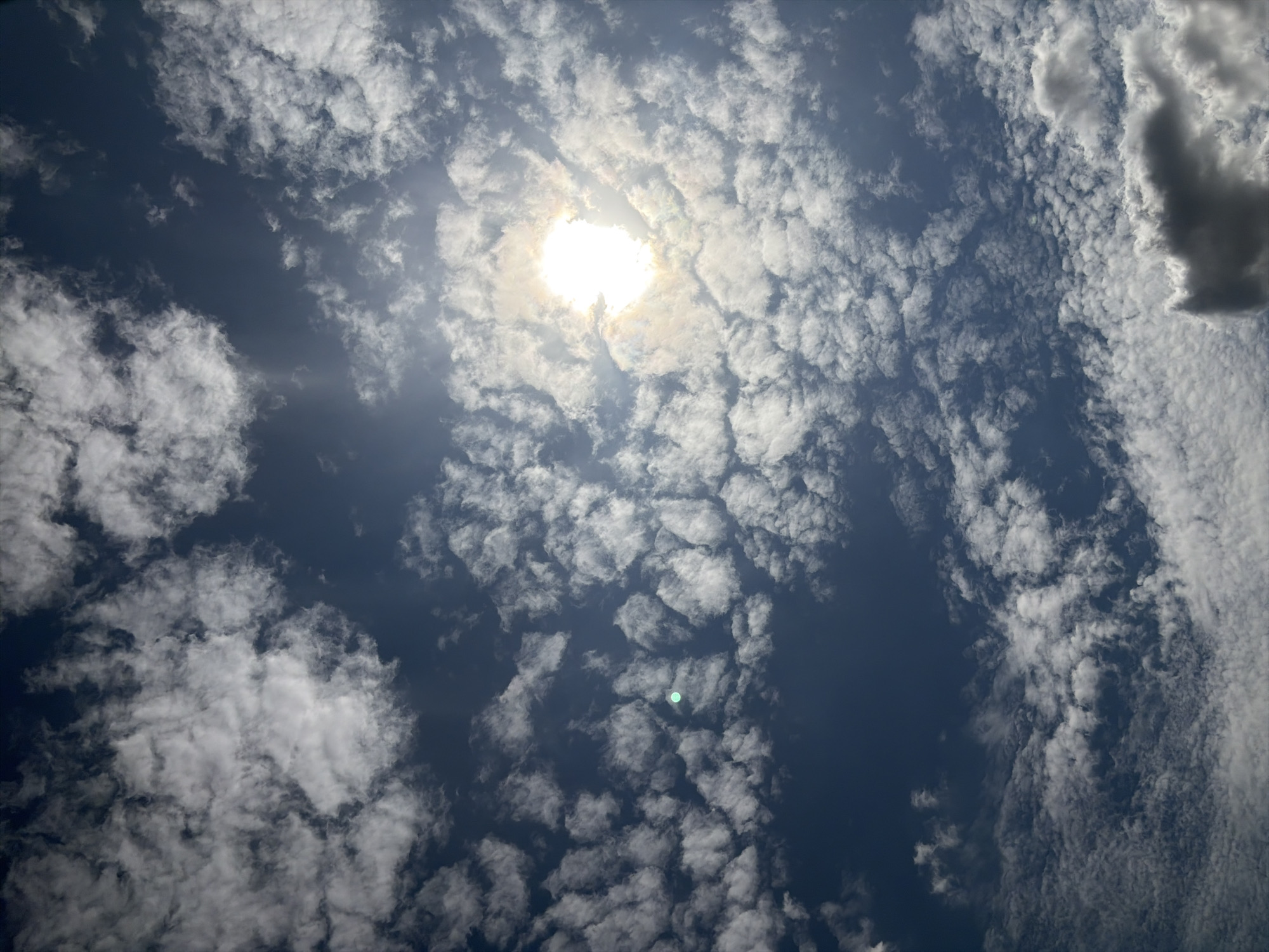 The image shows a sky filled with scattered white cumulus clouds against a deep blue sky. The sun is visible in the upper right quadrant, emitting a bright, diffused light through the clouds, creating a hazy effect. There are no people, objects, or buildings visible in the frame, as the image is focused entirely on the sky. There is a faint, circular, light green anomaly in the middle left of the image. The scene depicts a clear, partly cloudy day. The location is identified as Oxentea, Moldova.