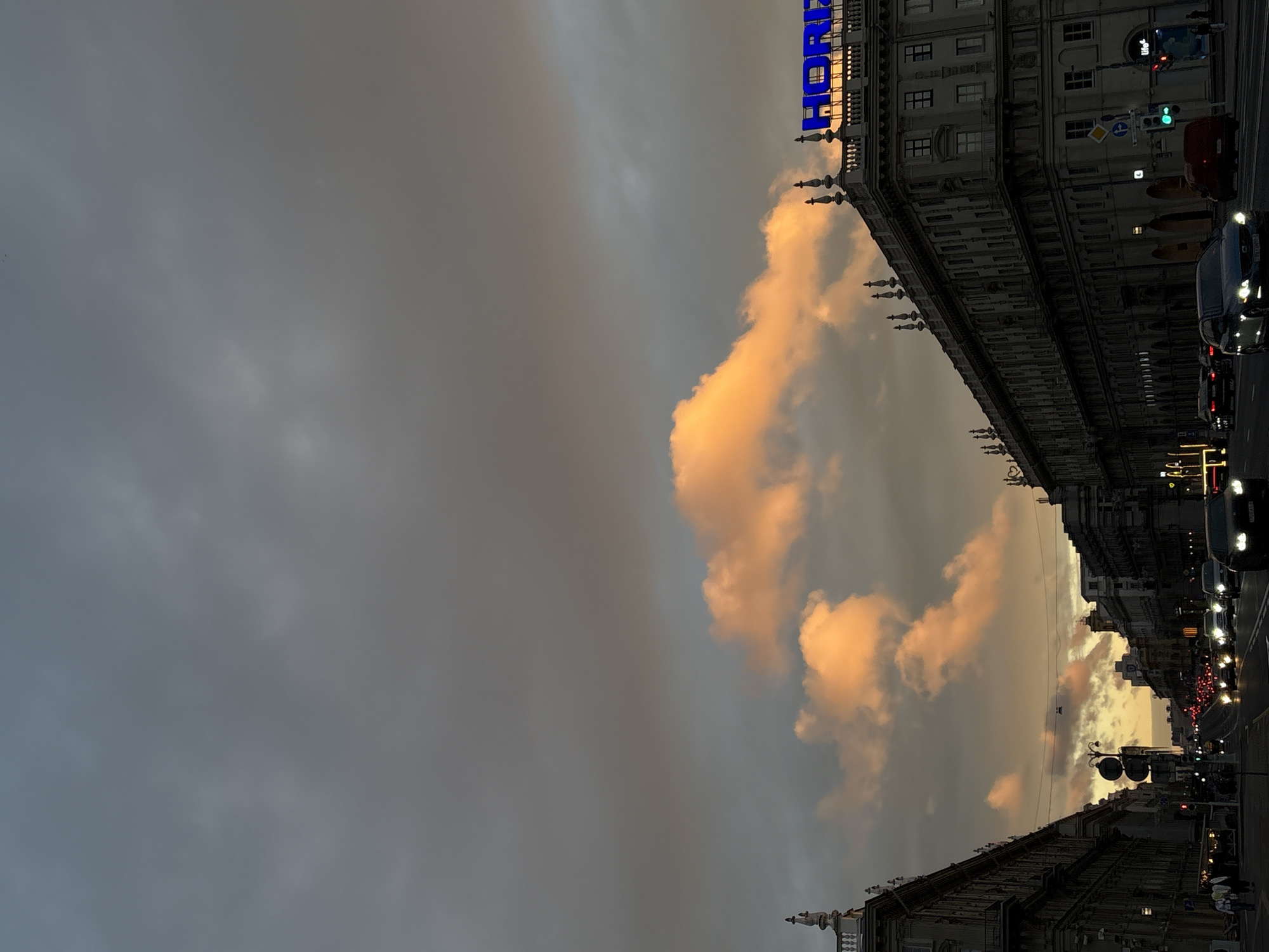 The image depicts a street scene in Minsk, Belarus, during twilight. The sky is dominated by dark, ominous clouds, with a band of bright, orange-tinged clouds cutting across the middle, likely illuminated by the setting sun. On the right side of the frame, a building with the illuminated blue sign "HORIZONT" is visible, characteristic of the HORIZONT department store on Nezavisimosti Avenue. The street below is lined with ornate architecture, typical of Stalinist Empire style buildings found along the avenue. A steady flow of cars, their headlights on, navigate the multi-lane road, indicating rush hour traffic. Pedestrians are sparsely visible on the sidewalks. The overall atmosphere is one of urban transition from day to night, with a dramatic sky adding to the scene.