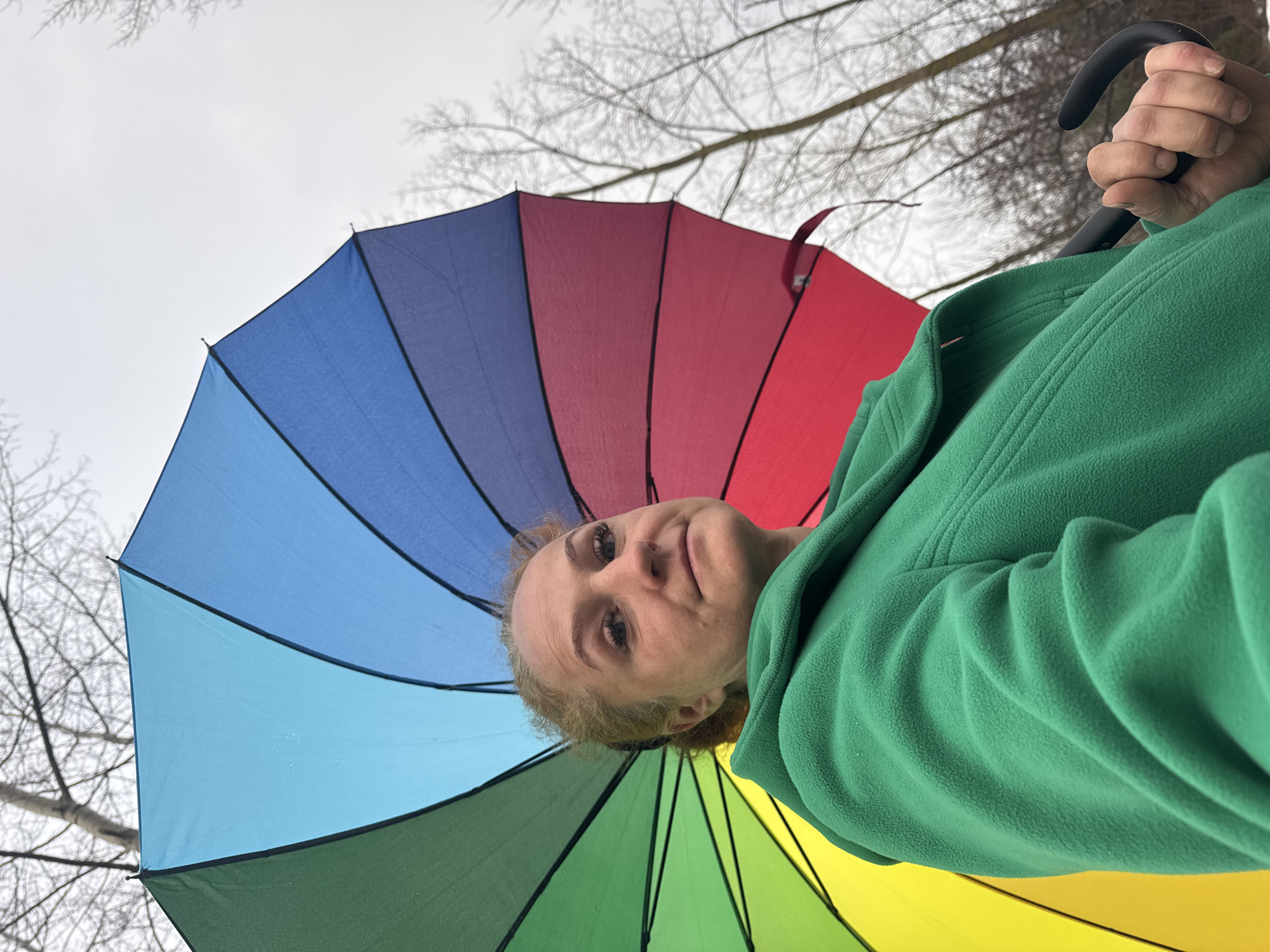 A low-angle, close-up shot shows a fair-skinned woman under a multi-colored umbrella in an outdoor setting. The umbrella features distinct sections in shades of blue, red, green, and yellow. The woman is wearing a green fleece jacket. Bare tree branches are visible against a gray sky in the background. The location is potentially the city of Chaikovsky, Russia.