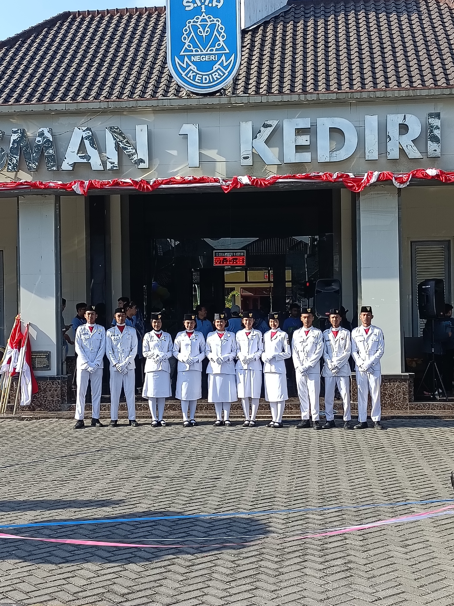 The image depicts the facade of SMA Negeri 1 Kediri, a school located in Kediri City, Indonesia. In front of the school entrance, a group of ten young people, dressed in matching white uniforms with black hats, stand in two rows. The individuals are a mix of males and females. The building's signage, "SMAN 1 KEDIRI," is prominently displayed above the entrance. Red and white streamers are draped across the front of the building, suggesting a festive or commemorative occasion. Several flags, also red and white, are planted to the left of the entrance. The ground in front of the school is paved with interlocking bricks, and two colored ribbons, one pink and one blue, are laid out on the ground. The environment appears to be outdoors, under daylight.