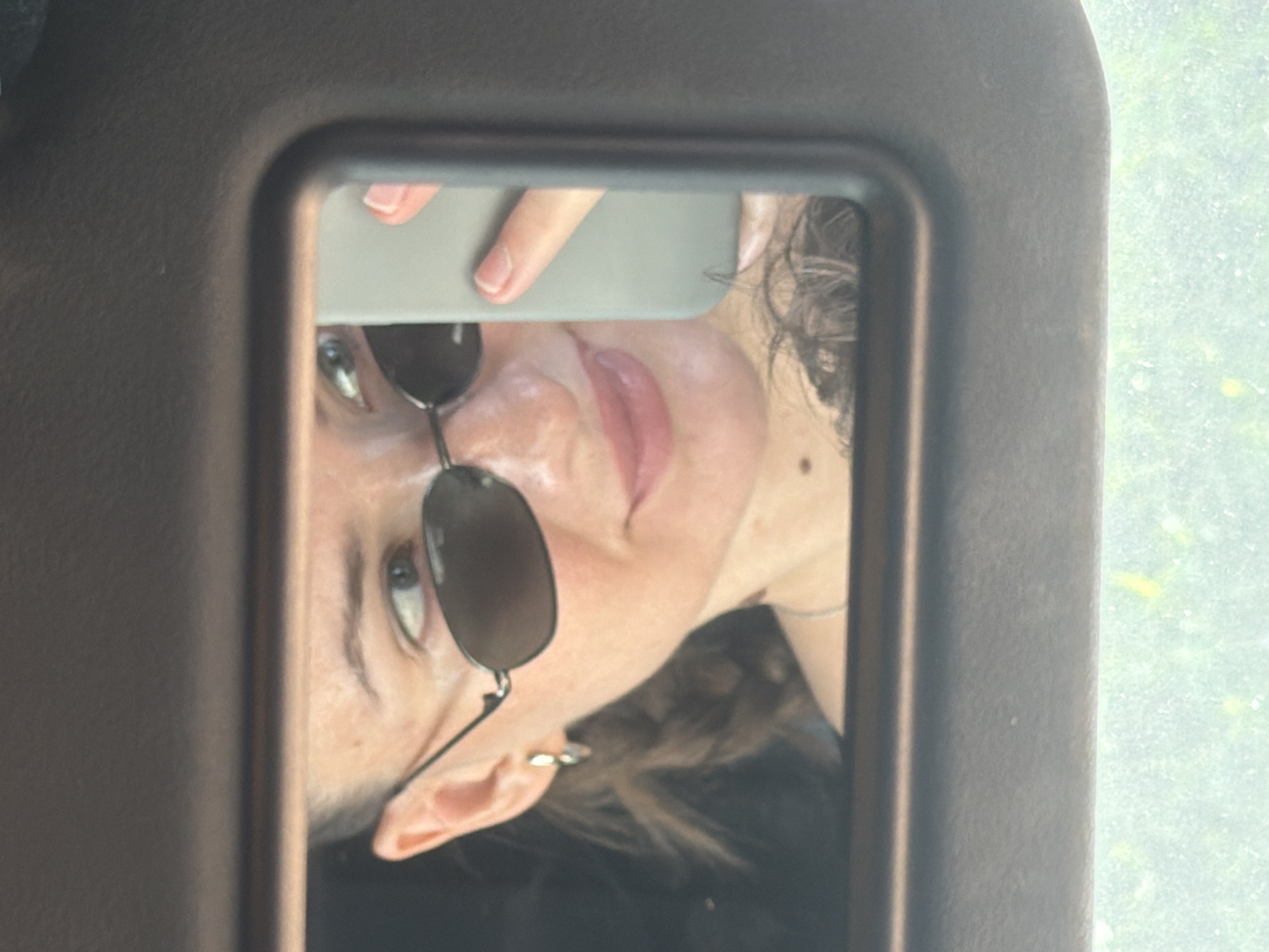 A woman wearing sunglasses and a silver hoop earring is reflected in a car's vanity mirror. She is holding a light grey smartphone, presumably to take the photo. Her hair is pulled back, and she has a mole on her chin. The background of the reflection shows a dark interior, possibly a car. The exterior visible below the mirror is out of focus, showing a blurry green and yellow pattern, suggestive of vegetation. This image was taken in Sharm El Sheikh, Egypt.