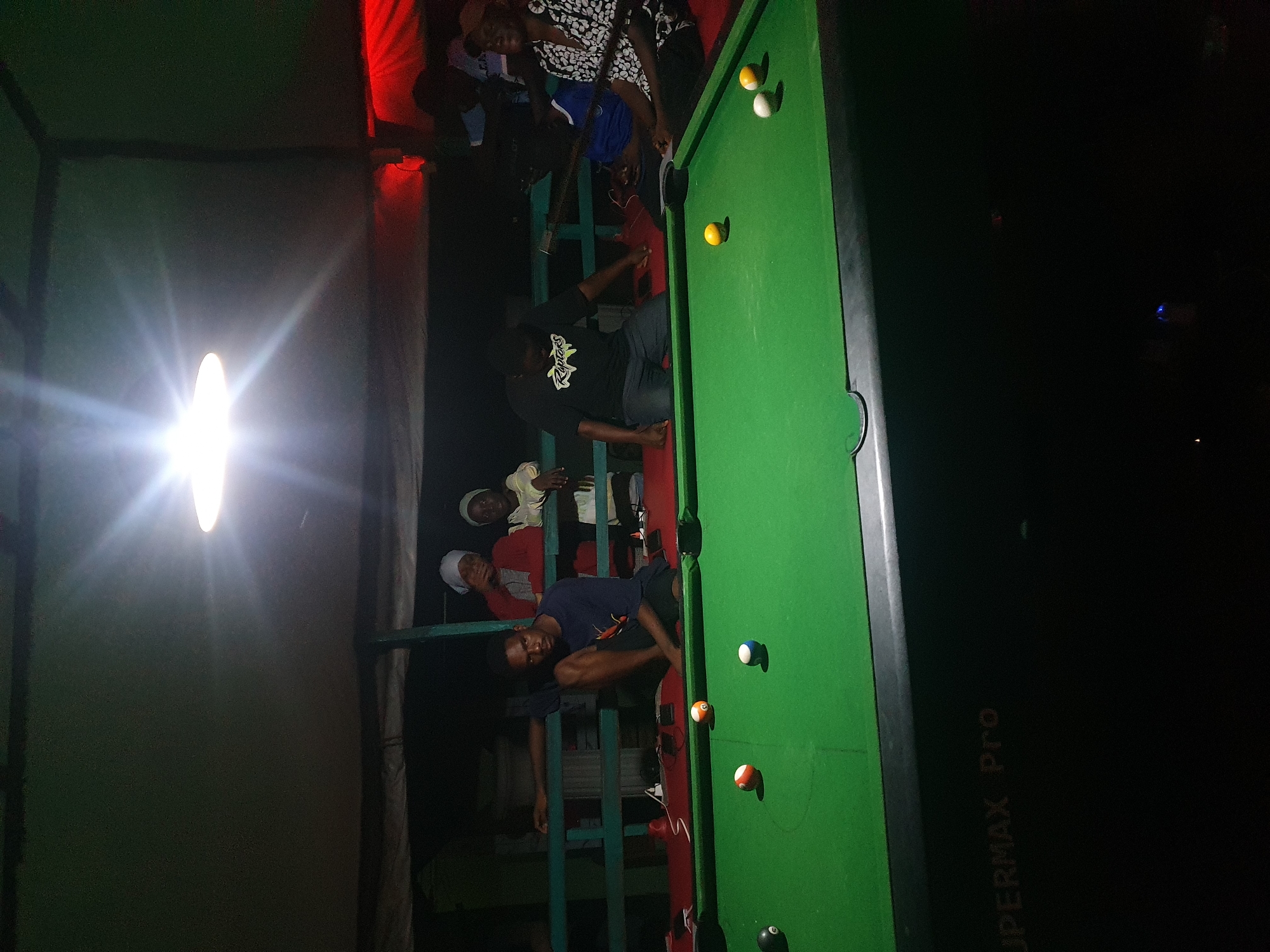 In Lokoja, Nigeria, a group of people gather around a pool table at night. The scene is illuminated by a bright overhead light, casting shadows and a warm glow. On the green felt pool table, several billiard balls are scattered, including a black ball, a striped orange and white ball, a solid white ball with a blue stripe, and a solid yellow ball. A man in a dark blue t-shirt is seated on the edge of the table, looking towards the camera. Behind him, two women in headscarves are standing, with another person in a red garment partially visible. To the right, a man in a black t-shirt with a "Raptors" logo is leaning over the table, holding a pool cue, and another person in a white and black patterned shirt is seated to his right. The background shows a dark, covered outdoor area with some reddish lighting in the distance. The pool table itself has the inscription "SUPERMAX Pro" visible on its side.