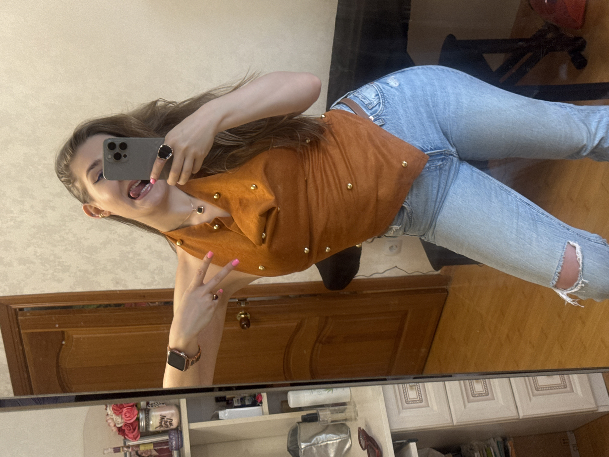 A young woman is taking a mirror selfie in a room. She is wearing a brown halter top with gold studs and light-wash ripped jeans. She has long brown hair, is sticking her tongue out, and is wearing rings and a necklace. On her left wrist, she is wearing a smartwatch with a rose gold band. To her left, a tall mirror reflects part of the room, including a wooden door and a shelf unit with various items. The room appears to be a bedroom or dressing area. The image does not contain any information about the city of Ussurijsk, Russia.