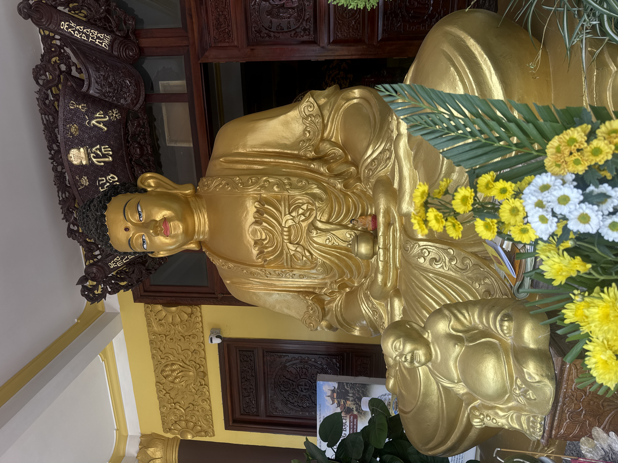 The image shows a large, golden Buddha statue seated in a cross-legged position. To the left and slightly in front of the main Buddha is a smaller, golden statue of a jovial, rotund figure, likely a representation of Budai or the Laughing Buddha. The statues are positioned in front of a building with yellow walls and dark wooden doors, adorned with intricate carvings. A wooden archway with carved dragons and what appear to be Vietnamese characters is situated above the Buddha's head. In the foreground, a bouquet of yellow and white chrysanthemums and green palm leaves is arranged. To the left of the statue, a brochure or book with an image of a temple can be seen. The overall scene suggests a serene, religious setting, likely within a temple or pagoda in Da Lat, Vietnam, given the architectural style and the commonality of such religious figures in Vietnamese culture.