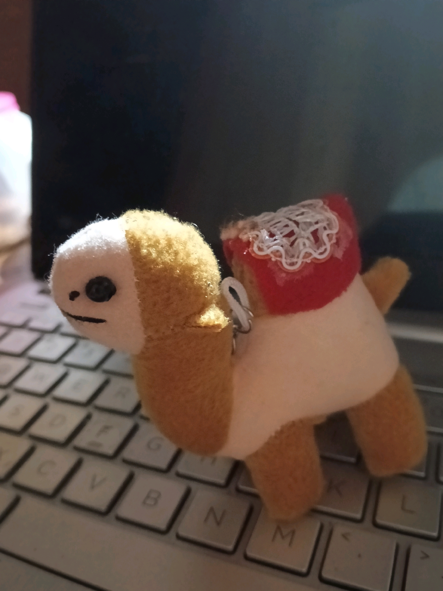A plush toy resembling a camel is positioned on a laptop keyboard. The toy is predominantly beige and white, with a small black eye and a black line for a mouth. A red fabric adorns its back, trimmed with white lace-like material. The keyboard shows individual keys with letters. In the background, out of focus, is a dark surface, possibly a laptop screen, and a blurry white object with a pink lid. The scene is indoors, with artificial lighting. There is no indication of the city of Galuk, Indonesia in the image.