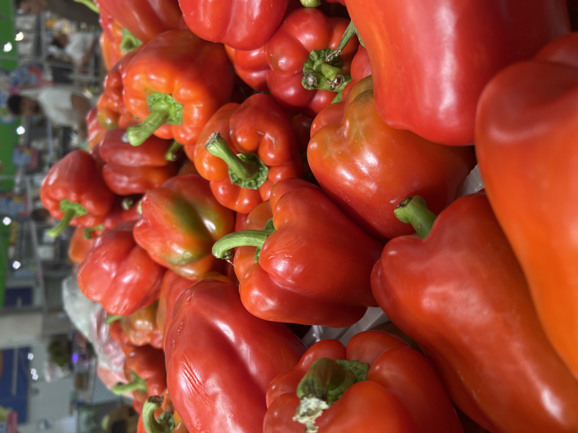The image shows a close-up, low-angle view of a pile of ripe red bell peppers at a market stall. The peppers are fresh and glossy, with some exhibiting a slight green hue near their stems. The background is blurred, revealing hints of a supermarket or grocery store environment with other produce and possibly shoppers in the distance. The lighting is bright, creating highlights on the peppers' surfaces. There is no indication that this scene is located in Yekaterinburg, Russia, or any other specific city.