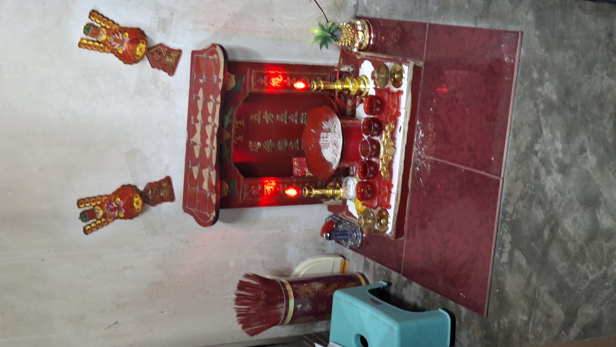 The image depicts an indoor shrine, possibly in a home in Lhokseumawe, Indonesia. The shrine is red and ornate, with a tiered roof. It features two red, flame-shaped electric candles, a gold-colored candle holder, and several small red cups arranged on a decorative red tray. Behind these items, within the shrine, are gold-colored characters on a red background. To the right of the shrine, a small, golden statue of a deity or effigy is visible, topped with a green, leafy plant. In front of the shrine, on a red tiled surface, are more red cups and decorative gold elements. To the left of the shrine, a tall, cylindrical container filled with red incense sticks stands next to a light blue plastic stool. A small lighter is also present on the shrine's platform. Two decorative paper cutouts, resembling ornate hands or figures with colorful embellishments, are affixed to the wall above the shrine. The wall behind the shrine appears to be a pale, textured surface, and the floor is a dark, concrete-like material. The overall scene suggests a religious or spiritual space.
