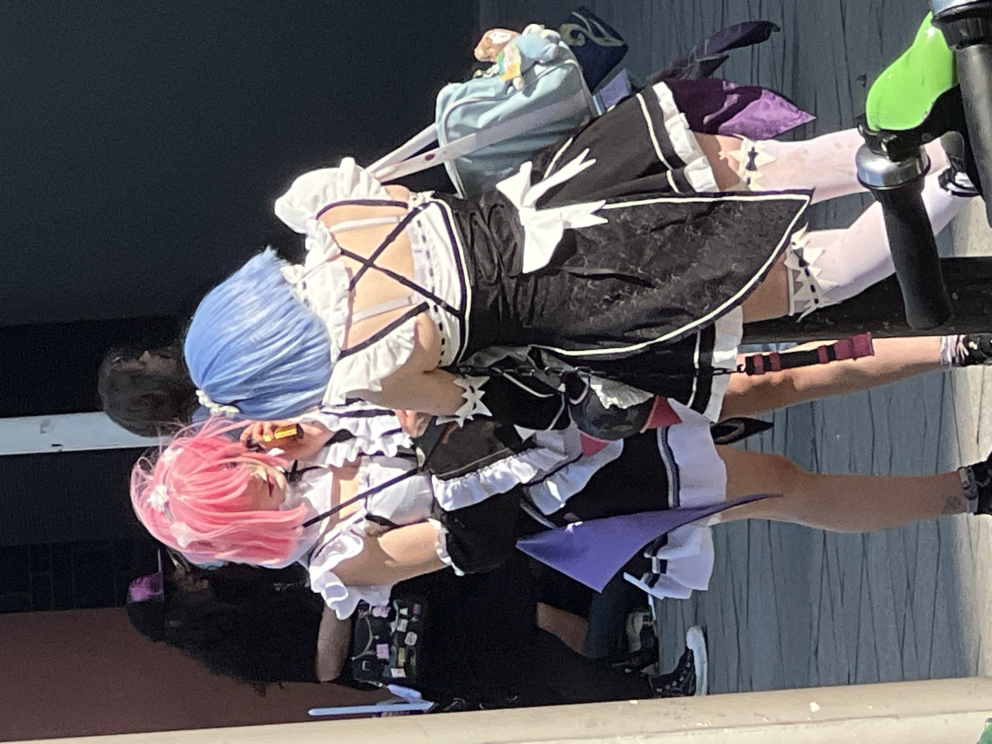 Two people dressed in anime cosplay outfits stand together. One has pink hair and the other has blue hair. Both are dressed as maid characters, wearing black and white frilly dresses, white thigh-high socks, and black shoes. They appear to be near a bicycle. The location is unknown.