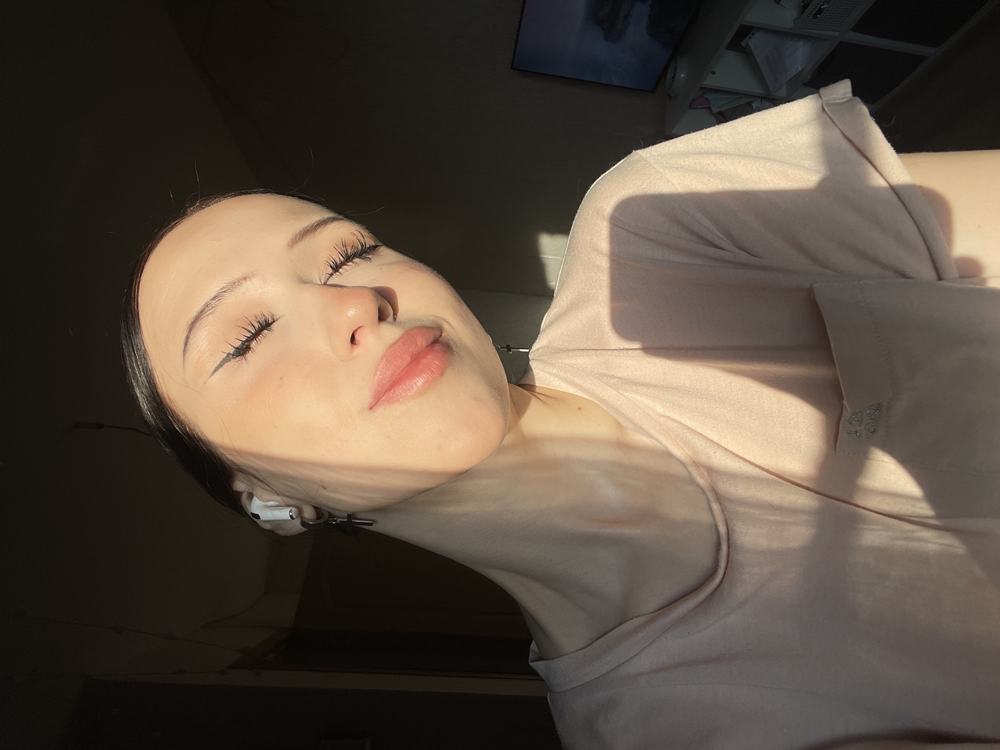This media file shows a close-up, side-profile shot of a young woman bathed in the warm light of what appears to be late afternoon sun. She is wearing a pale pink t-shirt. Her eyes are closed, and her head is tilted back, exposing her neck. She has on noticeable eye makeup, including winged eyeliner and long eyelashes, and her lips are painted with a glossy pink lipstick. She is also wearing white wireless earbuds. A small, dark cross earring is visible on her right ear. The background is largely dark, with some indistinct shapes suggesting an indoor setting, possibly a room with furniture and a television screen visible in the upper right corner. The strong sunlight casts distinct shadows across her face and clothing, highlighting the contours of her skin and the fabric of her shirt. The overall mood is serene and contemplative, with the lighting adding a dramatic and artistic quality to the image. The provided location context is Votkinsk, Russia. There is no visible text in the image.