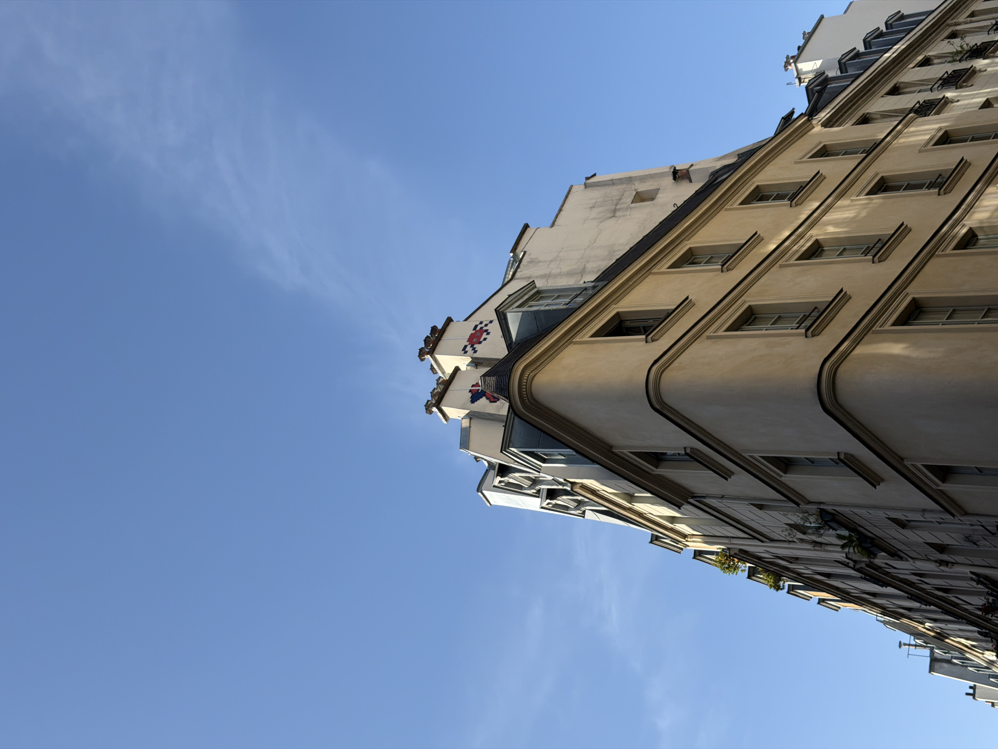 The image shows an upward-angled view of a Parisian building against a clear blue sky. The building features a series of windows with architectural moldings. At the top of the building, there is a decorative feature resembling a mosaic of red, white, and blue pixels. The overall aesthetic of the building is consistent with the architectural style found in Paris. The angle of the shot emphasizes the height and verticality of the building.