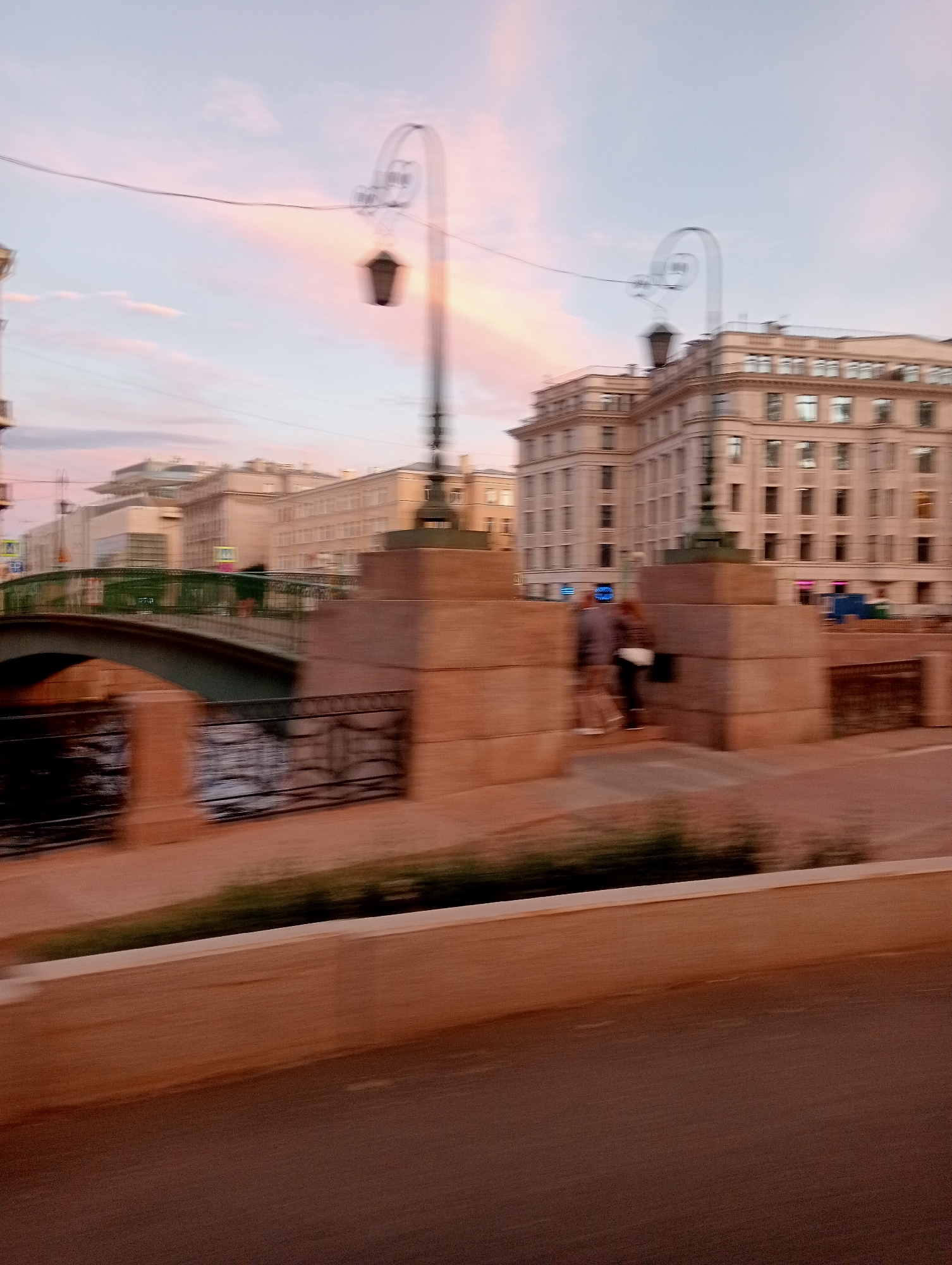 The image depicts a blurred view of a city street in Saint Petersburg, Russia, likely during dusk or dawn given the soft, pastel colors of the sky. The scene features a canal with a bridge crossing it, flanked by ornate street lamps and substantial stone structures. In the background, several multi-story buildings with classical architectural elements are visible. Two figures, also blurred, are present near the stone structures. The motion blur suggests the photograph was taken from a moving vehicle. The overall impression is of a serene, urban landscape.