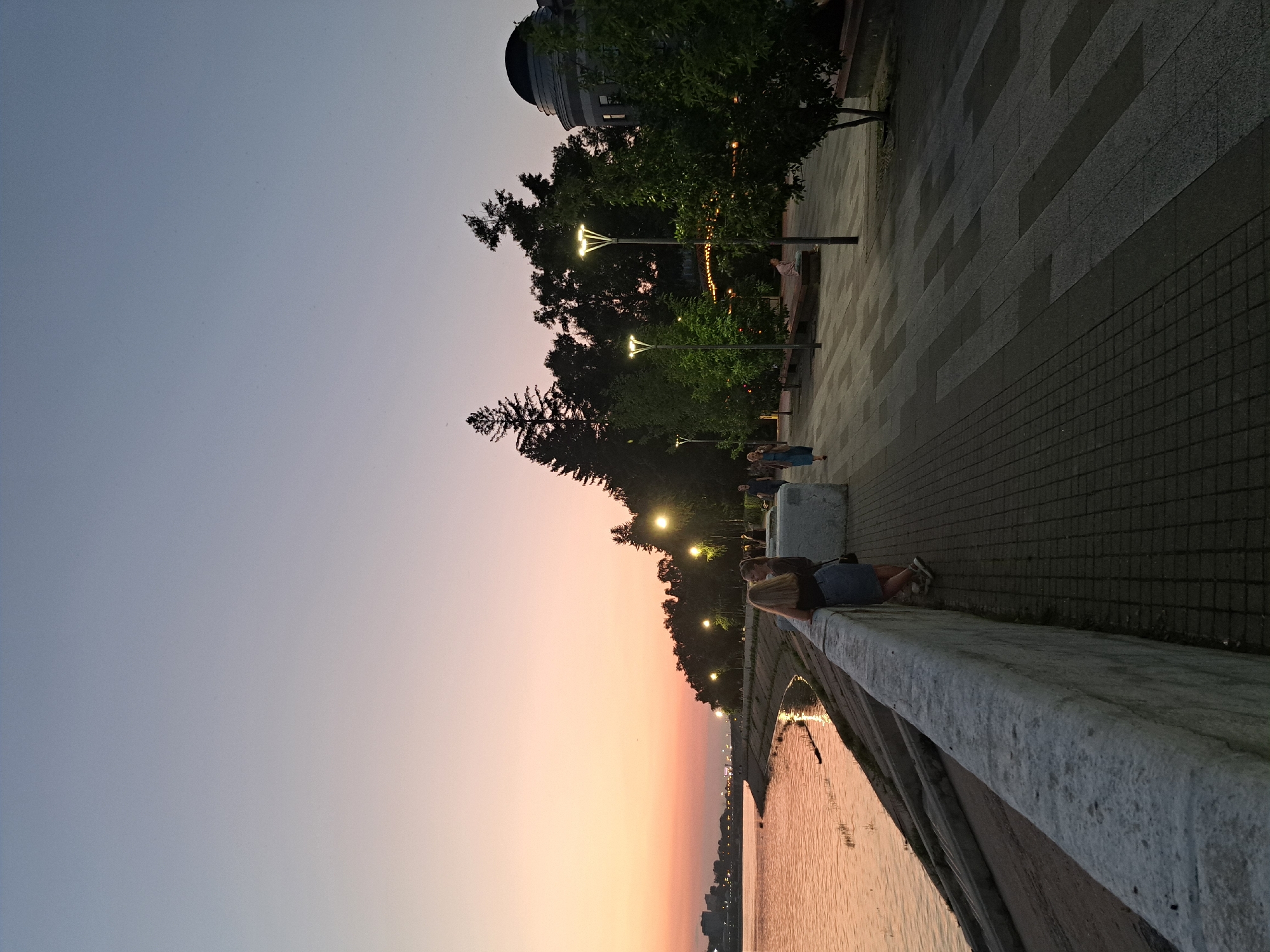 The image shows a waterfront promenade in Irkutsk, Russia, during twilight. A wide, calm river reflects the soft hues of the sunset, transitioning from pink to a pale blue at the horizon. The promenade is paved with dark and light grey rectangular tiles, illuminated by modern streetlights. Several trees line the promenade, casting shadows on the walkway. In the foreground, two young women stand near a thick, stone-block retaining wall, looking out towards the water. Further down the promenade, a few other individuals are visible, walking or sitting on benches. In the distance, the silhouette of a city skyline is discernible against the fading light. To the right, a domed building suggests the presence of significant architecture. The overall scene depicts a peaceful evening stroll along the riverbank.