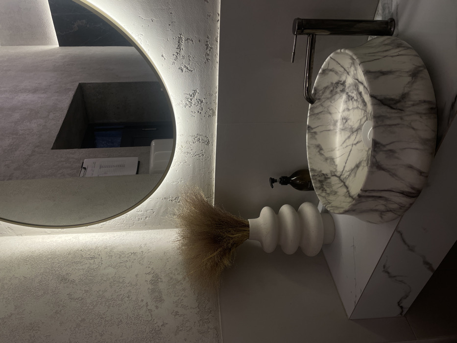 A modern bathroom features a round, backlit mirror reflecting a minimalist countertop. A unique marble sink sits beside the mirror, complemented by a dark metal faucet. A decorative white vase with dried grass rests beneath the sink. A dark brown soap dispenser adds a touch of elegance to the design. The color palette is predominantly white and gray, with accents of dark brown and metal.